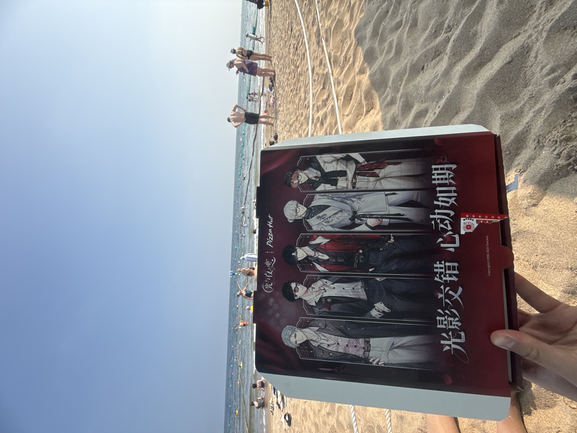 A person holds up a pizza box featuring animated characters on a sandy beach in Qinhuangdao, China. The pizza box has a dark red design with images of five men in stylish outfits and Chinese text. In the background, the blue ocean stretches towards the horizon, dotted with buoys and several people swimming and sunbathing. The sand is light brown and textured with ripples. The sky is clear and light blue.