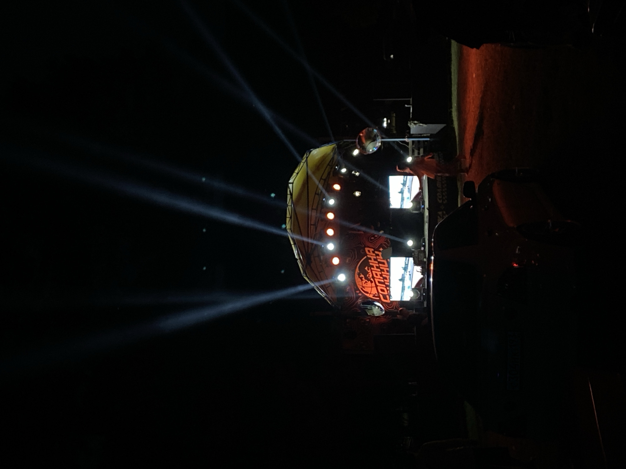 An outdoor nighttime festival or concert is taking place in Bezymyannoe, Russia. A large stage with a yellow canopy is illuminated by multiple bright white spotlights creating beams in the dark sky. A large digital screen is positioned on the stage, displaying what appears to be a racing simulation or video game. To the left of the screen, the word "СОЛНЦА" (Solntsa - Sun) is visible in large orange lettering on the stage's backdrop. There are several orange and white stage lights along the top of the stage structure. A person in an orange hooded garment is visible standing on the stage, facing away from the camera. In the foreground, a dark-colored car is parked, its license plate partially obscured by shadow. The ground appears to be covered in a reddish-brown dirt or gravel. Two large mirrored disco balls are suspended from poles on either side of the stage area, reflecting the light. Several other indistinct figures are visible in the background, partially silhouetted against the stage lights. The overall atmosphere is dark and festive, with a focus on the illuminated stage and performance area.