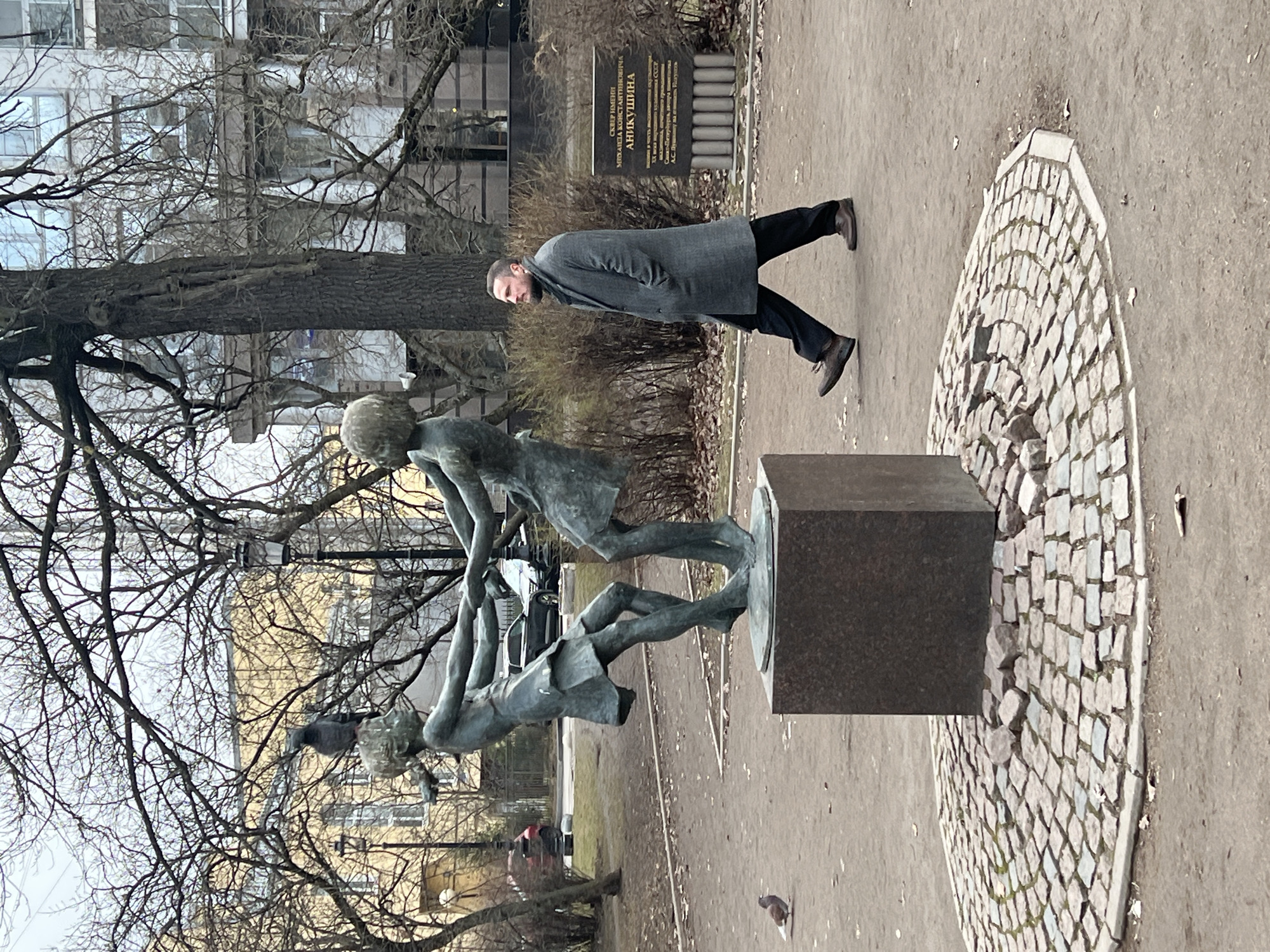 A sideways view of a public park in Saint Petersburg, Russia on an overcast day. A statue of three children playing is prominently featured, set upon a square stone pedestal surrounded by cobblestones and dirt. A man in a gray overcoat and dark pants walks to the right of the statue. A sign with Russian writing is visible in the background. Leafless trees dominate the skyline, framing the statue and building in the distance.