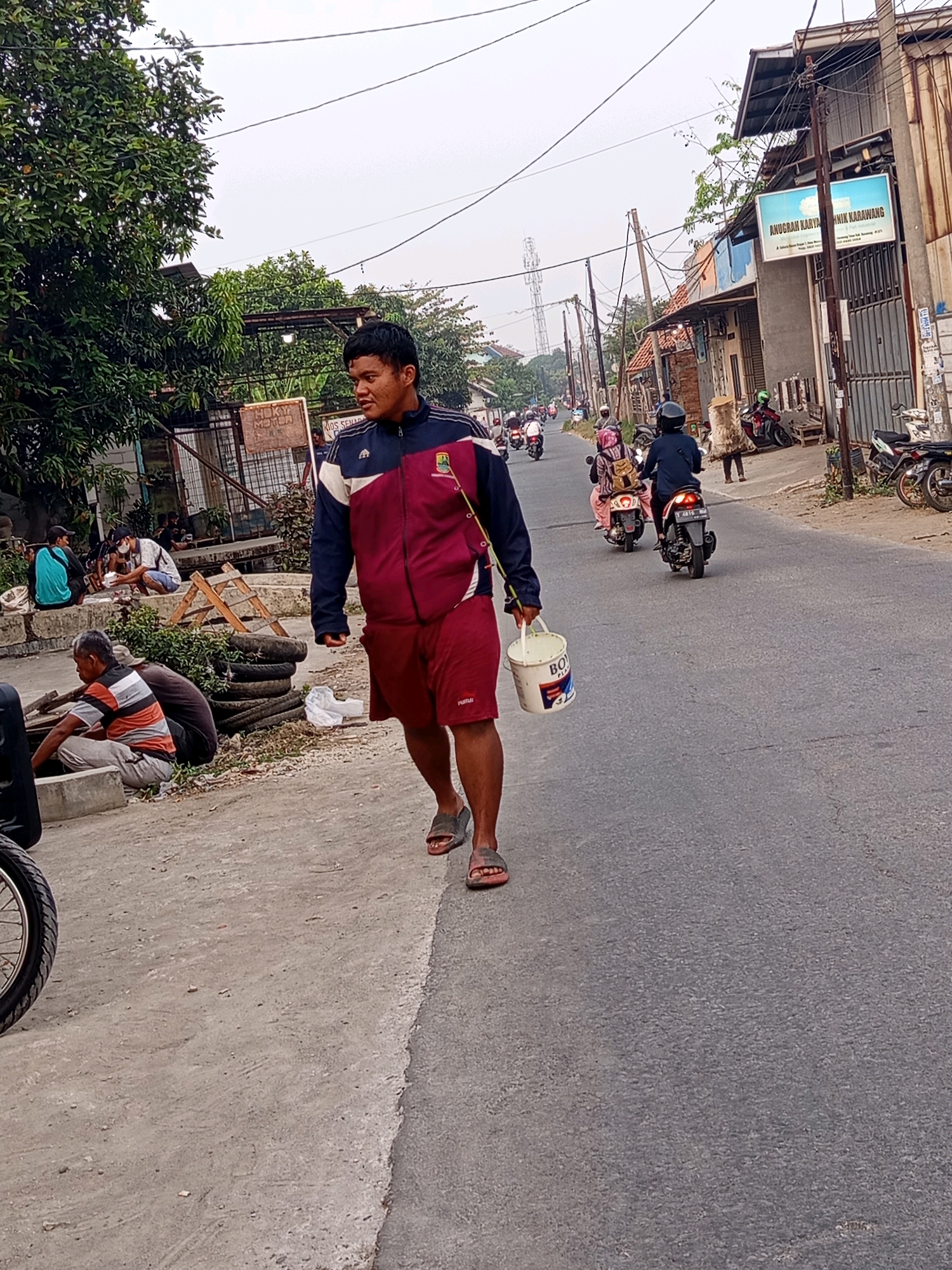 A full-shot, eye-level, outdoor vertical photograph captures a man walking along a paved street in Karawang, Indonesia. The man, who is the primary subject, is wearing a dark blue and maroon athletic jacket and maroon shorts. He is walking towards the right of the frame, carrying a white bucket in his right hand and a fishing rod over his left shoulder. His expression is neutral, and he appears to be walking at a moderate pace.
The street is lined with buildings on the right side, including a shop with a blue sign that reads "ANUGRCH KARYA TEKNIK KARAWANG". The buildings are made of various materials, including brick and corrugated metal. Several motorcycles are parked along the roadside, and others are visible on the street, with people riding them. A large tree with lush green leaves is on the left side of the frame, providing some shade. In the background, there is a utility pole with many wires. The sky is overcast, suggesting a potentially hazy or cloudy day.
On the left side of the street, there is a concrete embankment with several people sitting. Some are conversing, and others appear to be resting. There are also stacks of tires and some construction materials visible near the embankment. The overall scene depicts a typical street scene in a semi-urban or industrial area of Karawang, Indonesia.