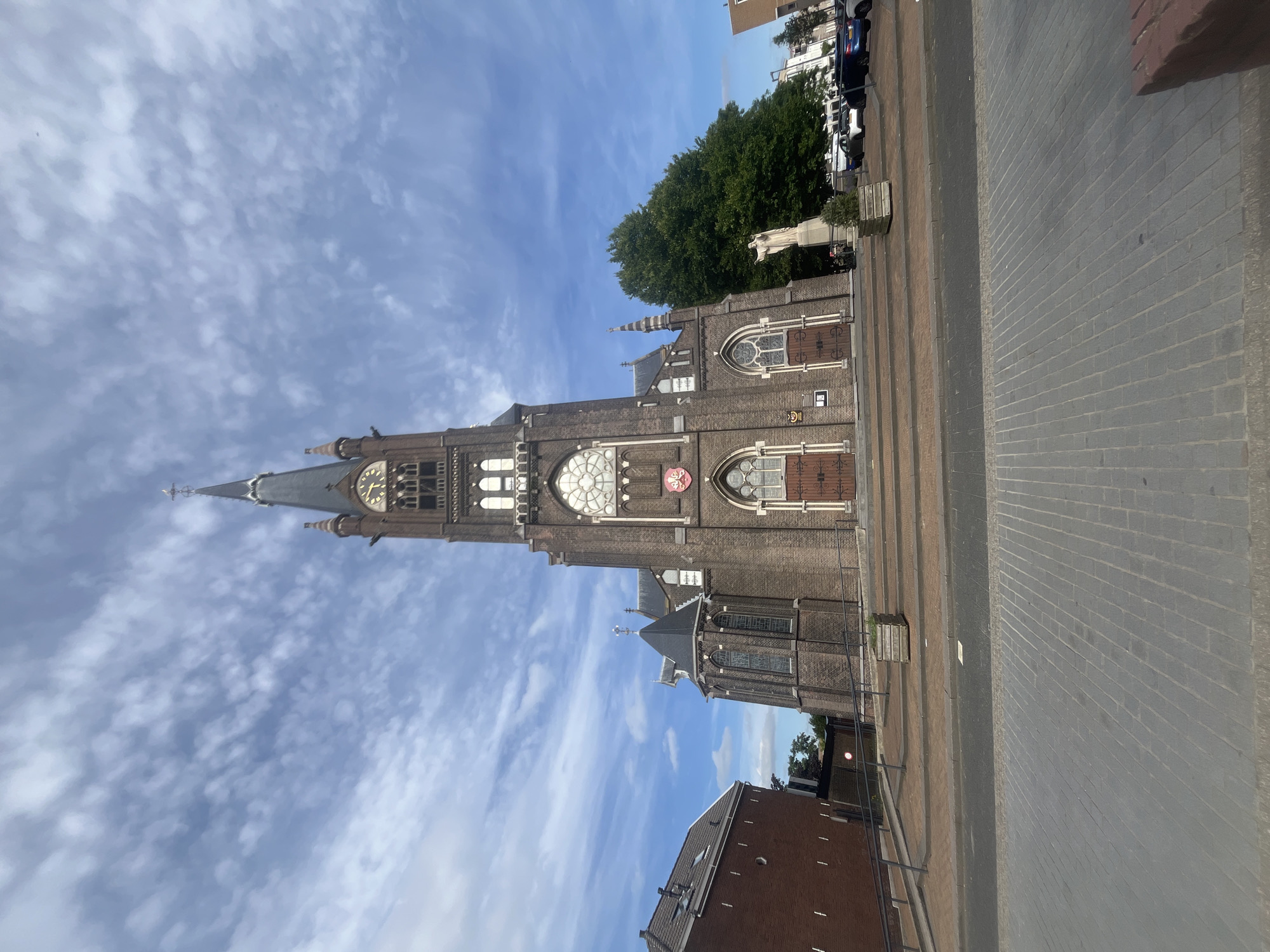 A wide-angle, low-angle shot captures the front facade of a large, historic brick church under a partly cloudy sky. The church features a tall, prominent steeple with a clock face and a cross at its apex. The building is constructed of dark brick and has arched windows and double wooden doors at the entrance. A shield with a red crest is visible above the main doors. To the left of the church, a smaller brick building with a pitched roof is visible. To the right, a large green tree stands next to a white statue of a standing figure. Cars are parked in the background. The foreground shows a paved area and a sidewalk. The overall scene depicts a view of the church from a public street.