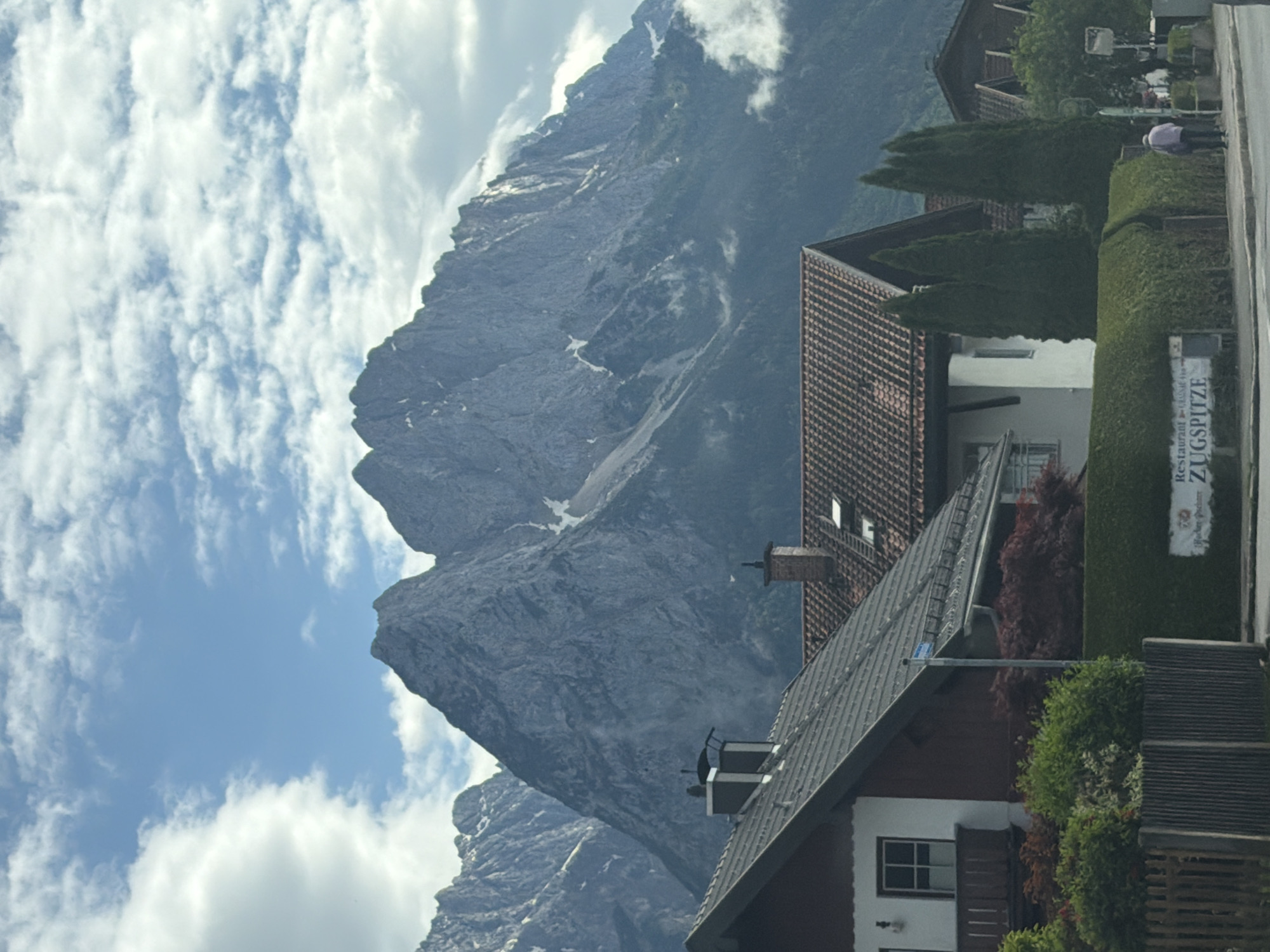 The image shows a mountain landscape, likely in a mountainous region, with a view of the Zugspitze peak. The sky is visible with scattered clouds. In the foreground, there are buildings with characteristic tiled roofs and some greenery. A sign with the words "Zugspitze" and "Restaurant" is visible. People are seen walking along a road or path. The location is likely in the city of Garmisch-Partenkirchen, Germany.