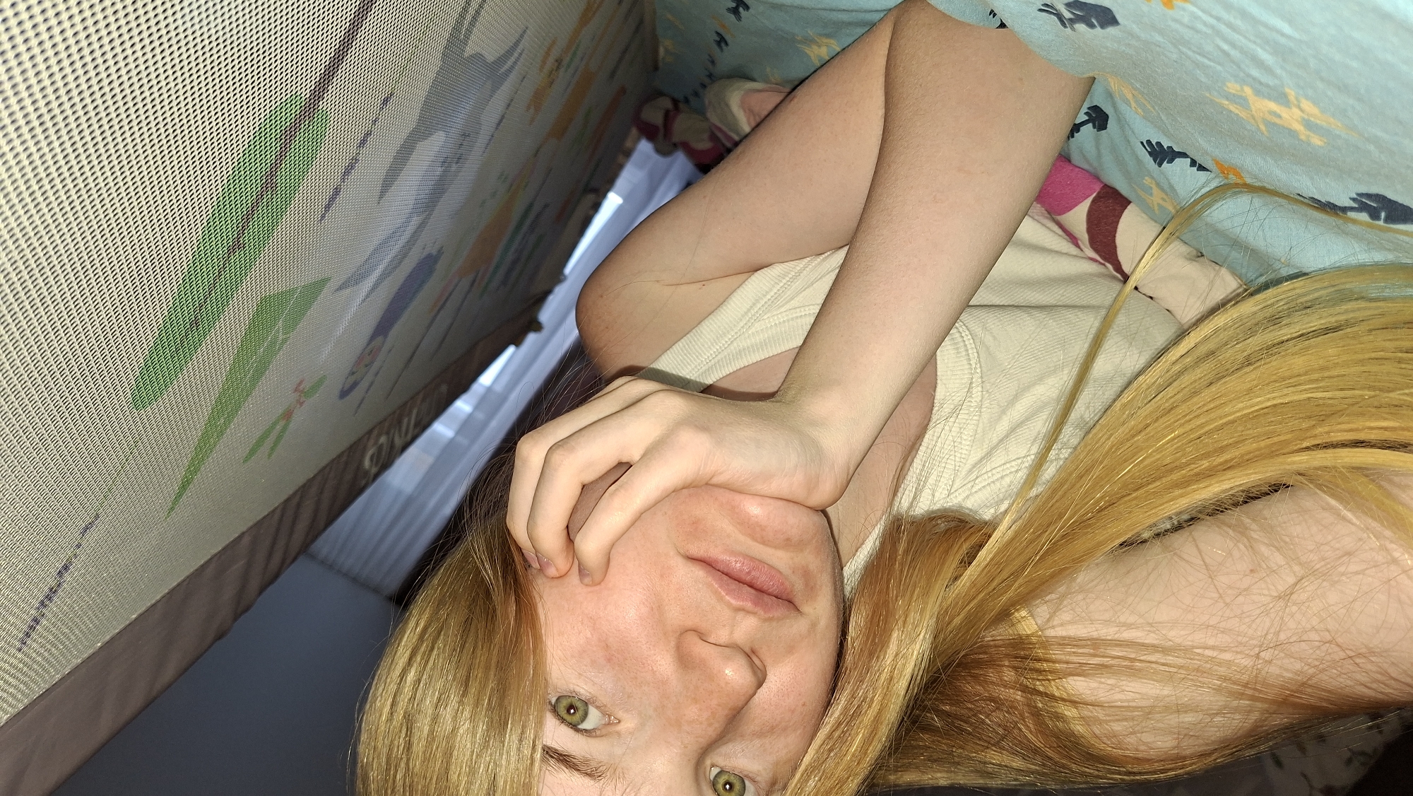 The image, rotated 90 degrees clockwise, features a person with fair skin, visible freckles, and long, straight blonde or light brown hair. The individual has green eyes and a neutral expression, looking directly at the camera. Their left hand is positioned near their chin. They are wearing a plain, light-colored (possibly off-white) top.
The setting appears to be indoors, likely a bedroom or play area. To the left of the person, a patterned, mesh-textured surface is visible, possibly part of a playmat, playpen, or bed rail. This surface displays various graphic shapes in green, blue, and other colors. Faint, partially visible text, difficult to decipher, can be seen along the bottom edge of this patterned surface. Beyond this, an out-of-focus window indicates an outdoor light source, suggesting it is daytime.
To the right of the person, a light blue blanket or duvet with abstract lighter-colored patterns (possibly yellow) covers part of the scene. Beneath the blanket, a pillow with pink and brown stripes is partially visible. The person's arms and upper body are seen against a light-colored sheet or fabric. The lighting is soft and natural. The person is in a state of rest, looking at the viewer.