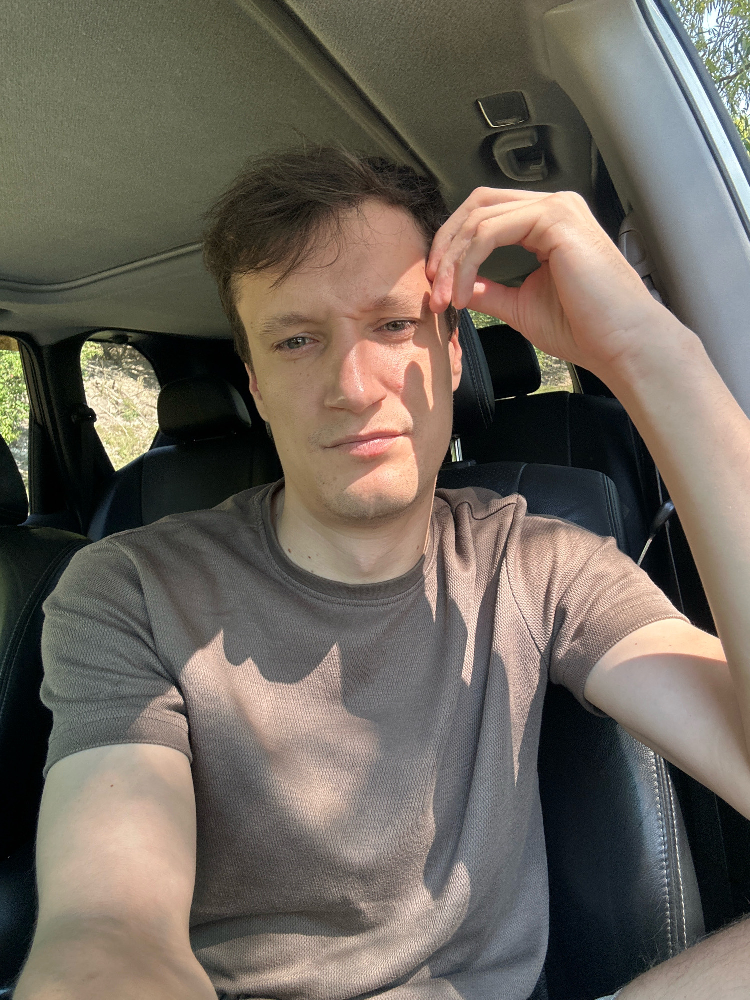A man is taking a selfie in a car. He is in the driver's seat, looking at the camera with his left hand resting on his forehead. He is wearing a brown t-shirt. The interior of the car is visible, including black leather seats and the car's ceiling. Sunlight is streaming in, casting shadows on the man and the car's interior. The background outside the car windows shows greenery and a bright sky. The location is not specified in Makiv, Ukraine, or any other city.