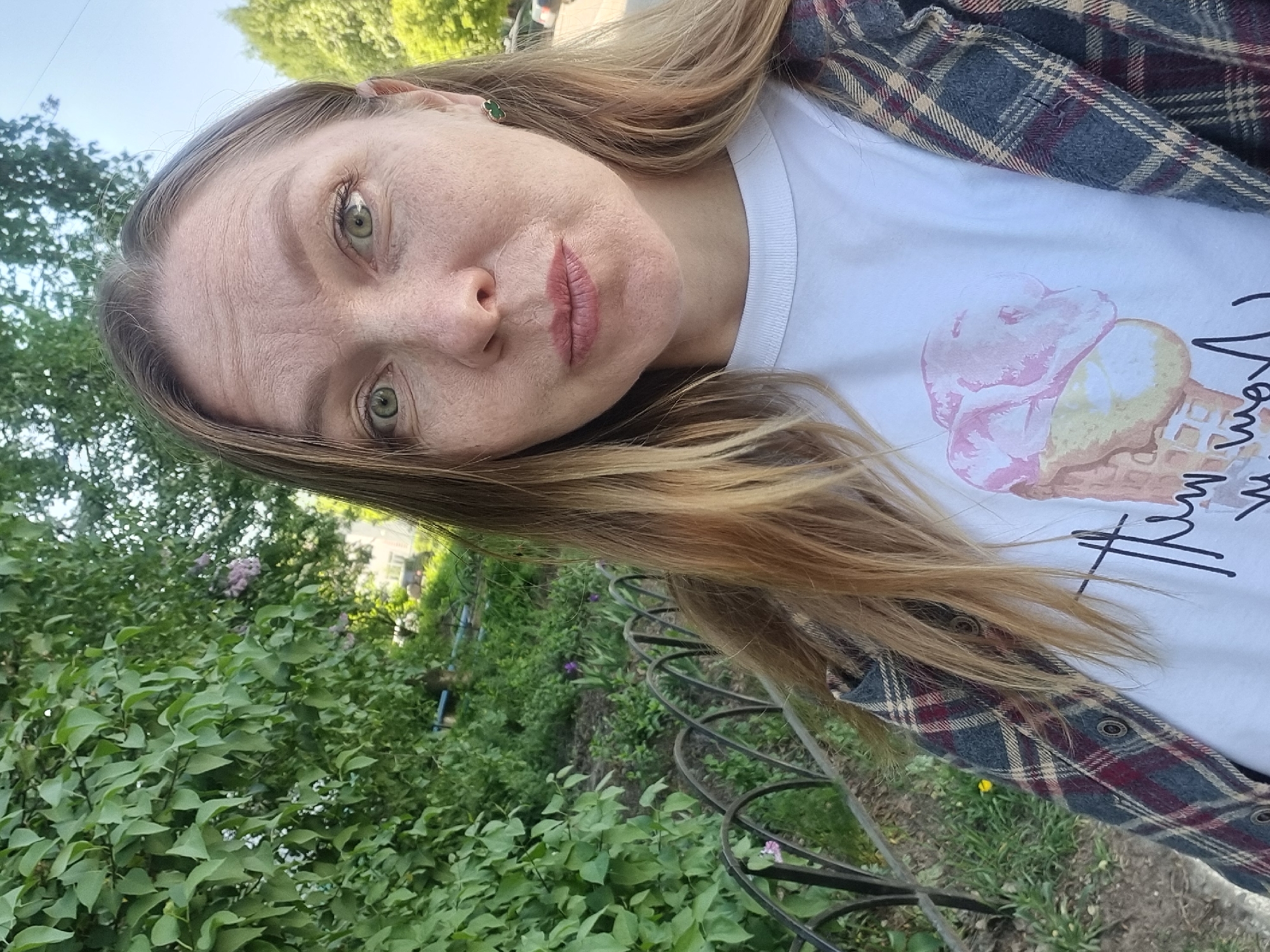 A woman with light brown hair and green eyes is posing for a selfie outside. She is wearing a white T-shirt with an ice cream cone design and a gray and burgundy plaid shirt. She appears to be standing in a park or garden, as there are green bushes and trees in the background. A black metal fence is also visible. The location is Chaikovsky, Russia.