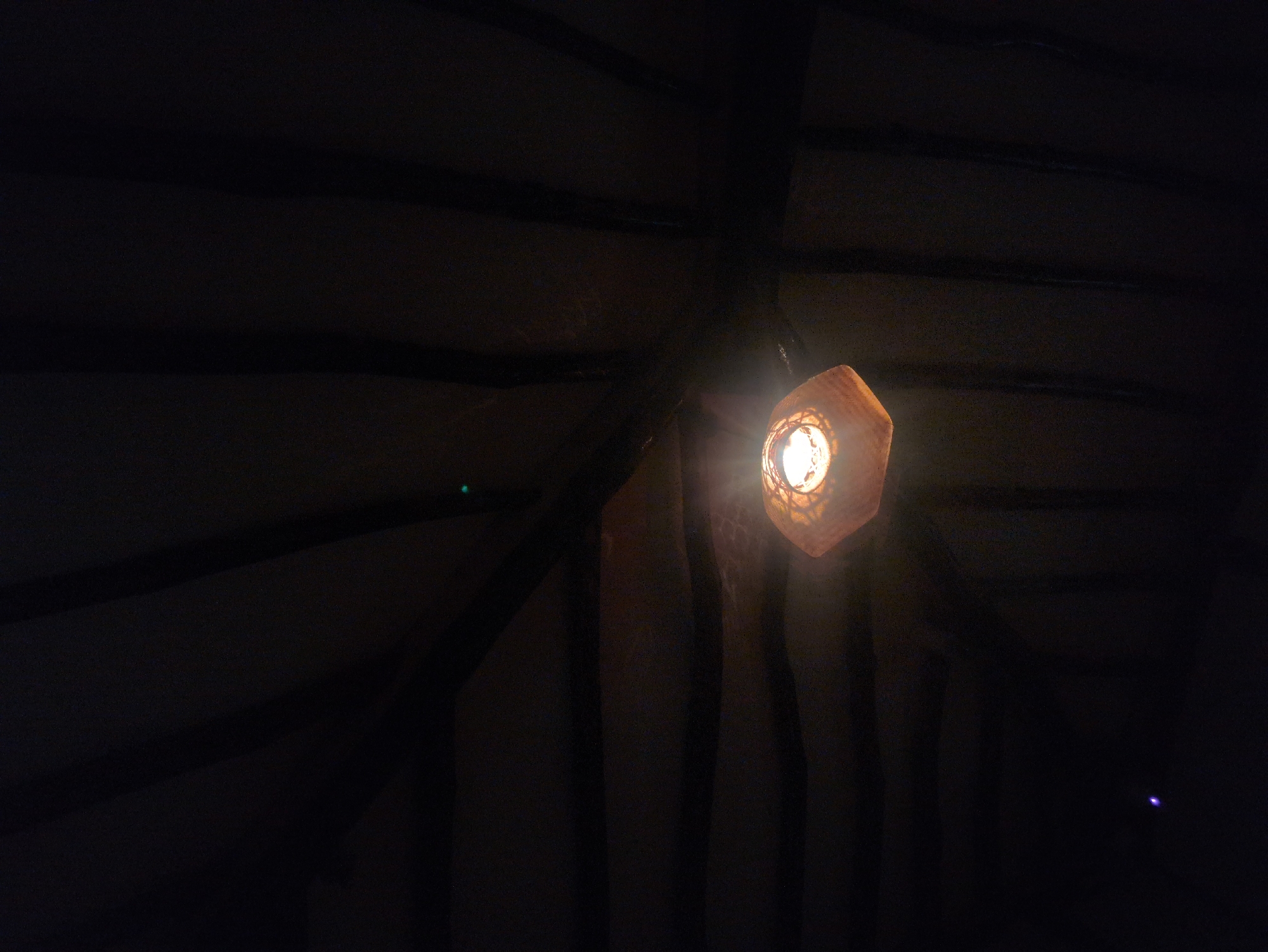 A dimly lit interior scene features a geometric, likely paper lantern hanging from the ceiling. The lantern's light illuminates the immediate surrounding area, revealing dark, horizontal ceiling beams. Dark vertical stripes, likely curtains or partitions, are partially visible below and to the side of the lantern. The location is unspecified and unidentifiable based on this image, making it impossible to confirm if it is in Seoul, South Korea. No people are visible.