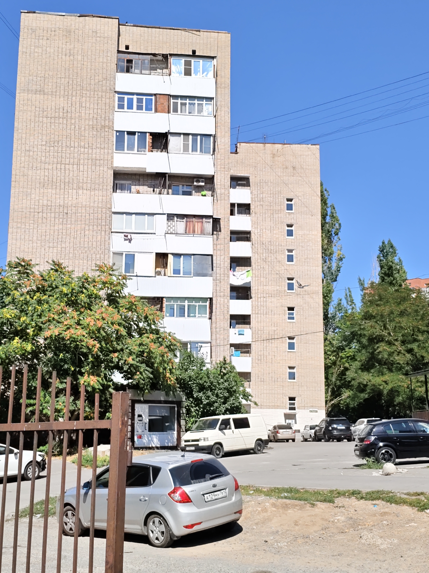 The image shows a tall, multi-story brick apartment building located in Rostov-on-Don, Russia. The building is light beige in color with white balconies and window frames. Several windows are visible, some with closed blinds or curtains. Various vehicles are parked in front of the building, including a silver hatchback in the foreground, a white van, and a black SUV. A metal fence is in the lower left of the image, with a gate slightly ajar. Trees with green and orange foliage are growing around the building, and a clear blue sky is visible above. A small kiosk with a glass front is situated near the fence. The area appears to be a residential courtyard or parking lot.