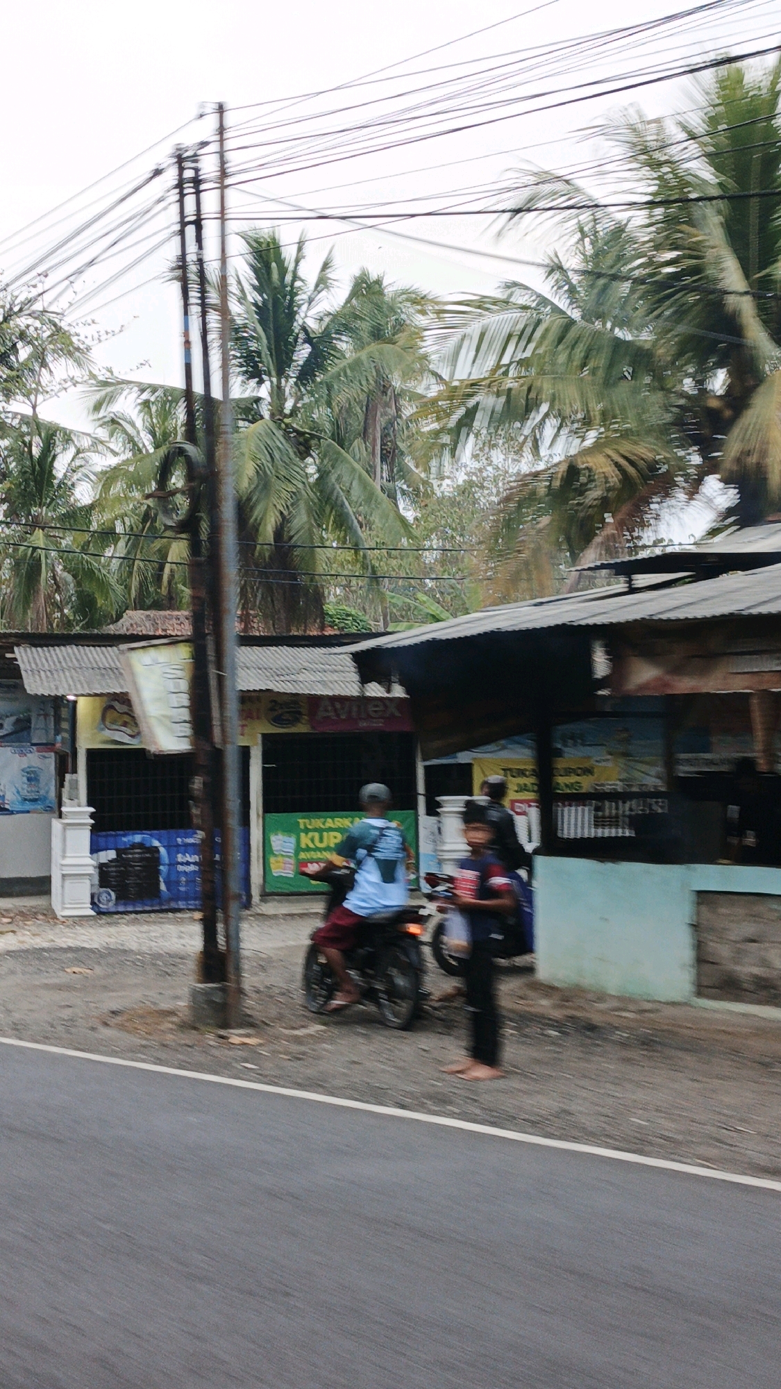 This image captures a street scene in Sukaresik, Indonesia, characterized by a slightly overcast sky and lush palm trees lining the background. In the foreground, a paved road runs diagonally across the frame. On the right side of the road, a row of small shops or stalls with corrugated metal roofs is visible. One stall prominently displays the "AvilEx" logo.
In front of the shops, several people and a motorcycle are present. A person is riding a motorcycle, wearing a blue shirt and red shorts, with their feet on the footpegs. Another person is standing to the right of the motorcycle, wearing a dark shirt and pants, barefoot. The ground in front of the shops is covered with gravel and dirt. Electrical wires are strung overhead, supported by a utility pole located near the center of the image, slightly to the left. Various signs and banners are affixed to the shops, some with Indonesian text, indicating commercial activity. The overall impression is one of a casual, everyday scene in a tropical urban or semi-urban environment.