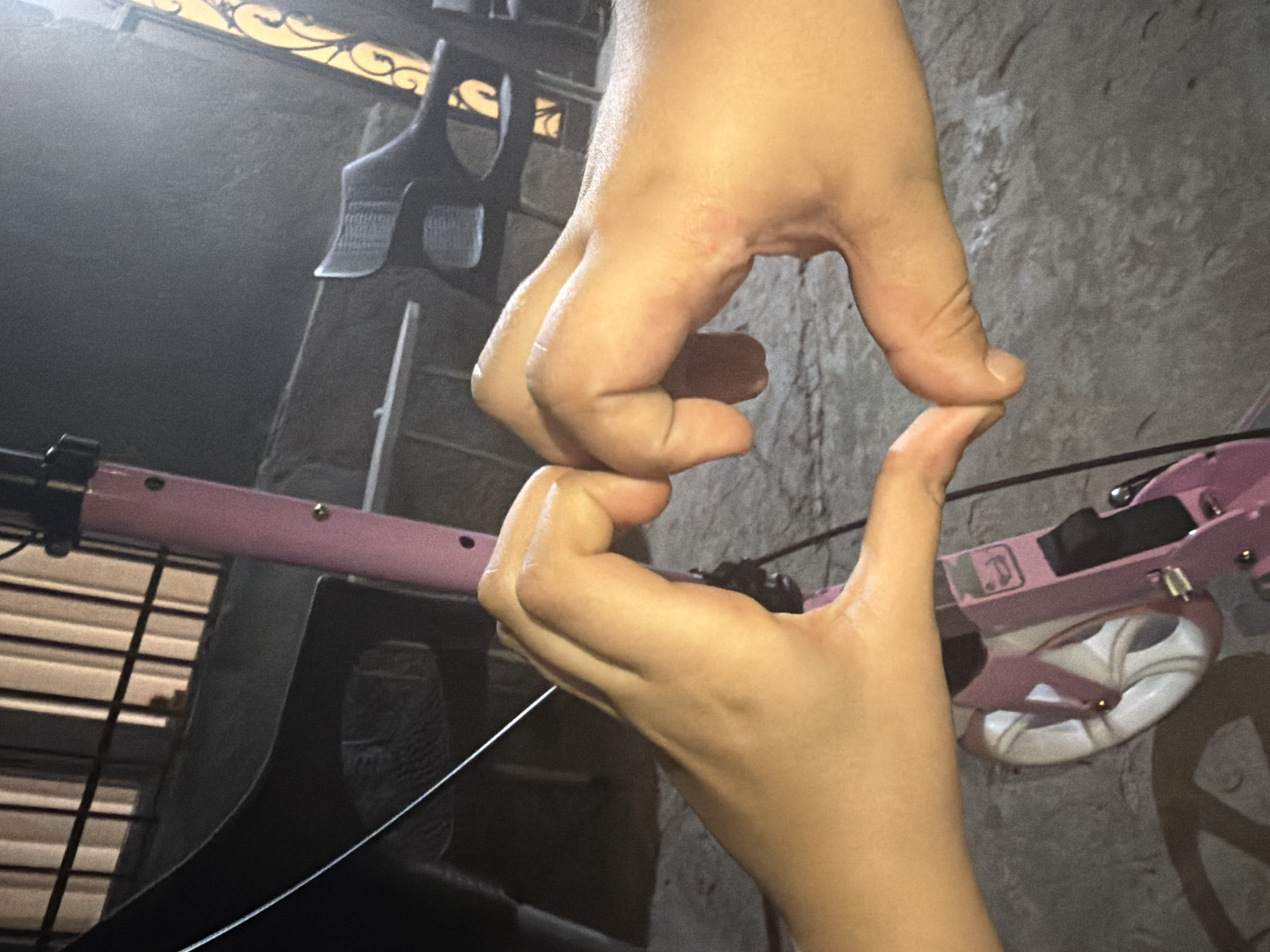 Two hands, likely belonging to a child, are prominently featured in the foreground, positioned to form a heart shape with their fingers touching. The skin tone is light.
In the blurred background, a pink scooter is visible, with parts of its handlebar, deck, and one white wheel with spokes. Behind the scooter, a dark, textured surface, possibly a wall or ground, fills much of the scene. To the left, a black object resembling a stool or chair with a patterned surface is present, along with vertical elements that could be part of a ladder or another structure. Above, a section of an ornate, dark metal railing or gate with golden accents is visible.
The lighting in the image is dim, suggesting it was taken during the evening or at night. The overall impression is one of affection or playfulness, conveyed by the heart gesture. No discernible text is visible within the image. The scene is set in Yerevan, Armenia.