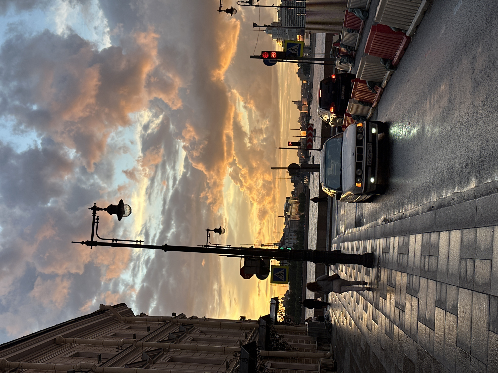 This image depicts a street scene in Saint Petersburg, Russia, during what appears to be sunset or sunrise. The sky is filled with dramatic clouds painted in hues of orange, yellow, and gray, suggesting the golden hour. A streetlamp with a classic design stands silhouetted against the colorful sky, casting long shadows on the paved sidewalk.
The scene includes several people walking along the sidewalk, their figures elongated by the low-angled light. One person is wearing a light-colored jacket and pants. The street itself is wet, reflecting the streetlights and headlights of passing cars, indicating recent rain or a damp atmosphere.
A light-colored car with its headlights on is prominently featured on the right side of the road. Another darker car is visible further down the street. The road is bordered by construction barriers, suggesting ongoing work or maintenance. In the background, the silhouette of the city skyline can be seen across a body of water, with various buildings and structures. A traffic light is visible, showing a red signal. A pedestrian crossing sign is also present.
The overall mood of the image is serene and atmospheric, with the dramatic lighting and the quiet presence of pedestrians creating a contemplative feeling. The presence of architectural elements like the ornate building facade on the left and the streetlamp suggest a historic urban environment. The license plate of the car is partially visible, reading "7554MM 198".