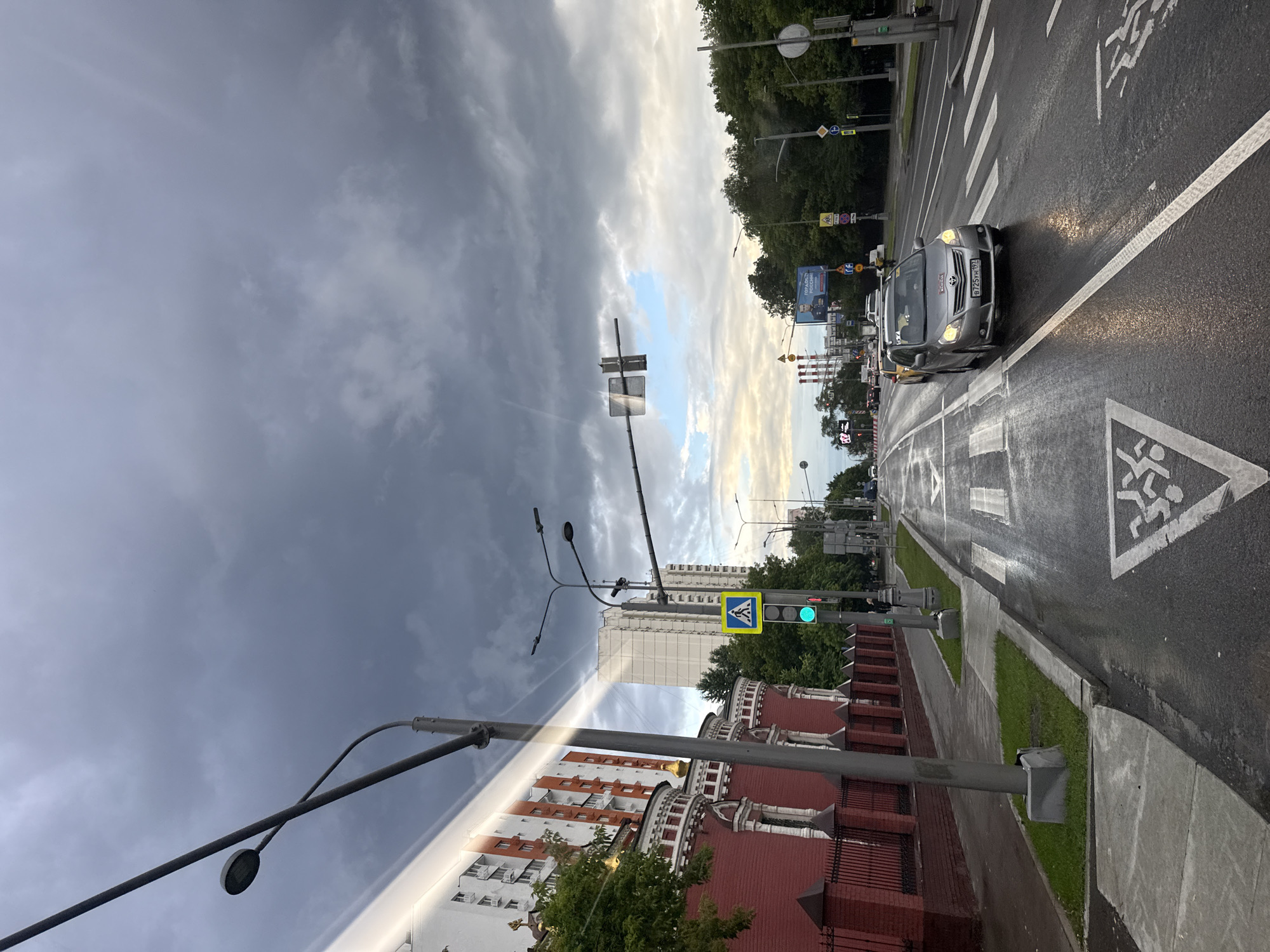 This is a vertical, full-shot photograph of a street scene in Moscow, Russia. The image is split horizontally, with the top half dominated by a dramatic, cloudy sky and the bottom half depicting the street and buildings.
The sky displays a mix of dark, stormy clouds on the left and lighter, brighter clouds with hints of blue sky on the right, suggesting a change in weather or a sunset/sunrise.
On the right side of the image, a wet asphalt road with white pedestrian crossings is visible. A silver car is driving down the road, its headlights on. Traffic lights, street signs, and lampposts line the street. Green pedestrian crossing lights are illuminated.
On the left side, a red brick building with white trim and arched windows is partially visible, suggesting traditional Russian architecture. Trees and tall apartment buildings can be seen in the background. The wet road surface reflects the overcast sky, contributing to the overall moody atmosphere of the image.
There is some visible text on a billboard in the background, but it is too blurry to read. The license plate of the car is visible and reads "B725MK 192". The overall impression is a moment captured on a potentially rainy day in an urban environment, with a stark contrast between the stormy sky and the urban landscape below.