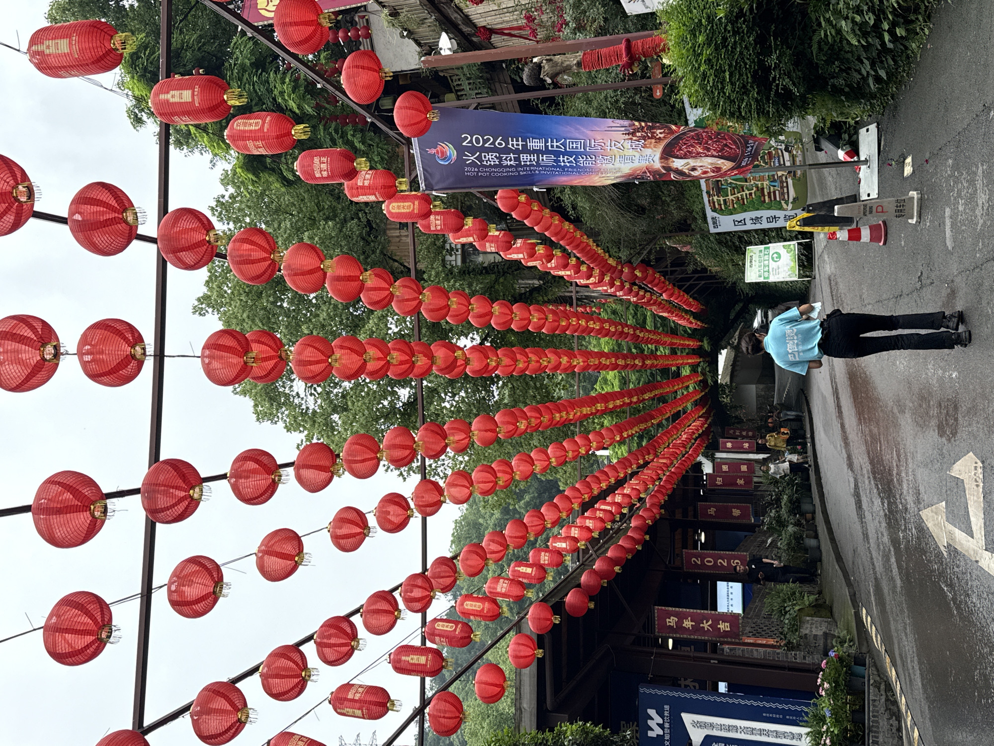 A slightly rotated, high-angle shot depicts a street in Nan'an District, China, adorned with rows of red lanterns strung overhead, creating a tunnel-like effect. The lanterns vary in shape, some elongated and cylindrical, others spherical. Many of the cylindrical lanterns have Chinese characters printed on them.
A banner hangs above the street, featuring the text "2026 Chongqing International Friendship Hot Pot Cooking Skills Invitation Competition" in English and Chinese. Various signs and advertisements flank the street. The signs are in Chinese, with text like "Welcome" and "Ma Nian Da Ji" (Happy Year of the Horse).
A person wearing a light blue shirt and dark pants stands on the right side of the image, facing away from the camera. They appear to be either reading or writing on something. The street surface is wet, reflecting light and adding to the diffused ambiance of the scene. Trees are visible beyond the lanterns. Traffic cones and parking signs are also present along the street.