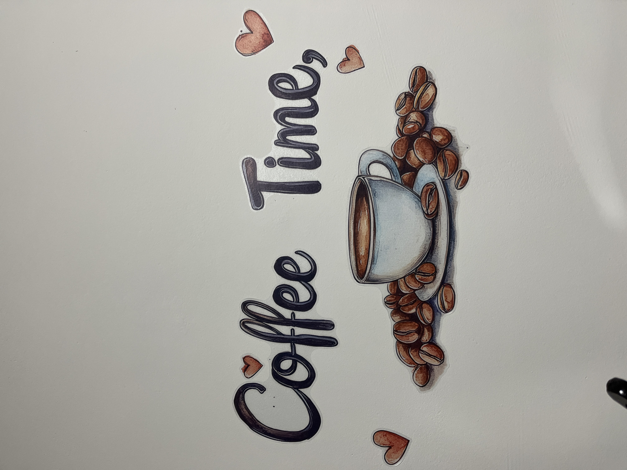 The image shows a decorative sticker on a light-colored wall, likely in a coffee shop or cafe in Garmisch-Partenkirchen, Germany. The sticker depicts the words "Coffee Time" written in stylized, dark lettering with a gradient effect. Several small, red heart shapes are placed around the text. Below the text is an illustration of a white coffee cup filled with dark liquid, resting on a saucer. A pile of coffee beans surrounds the cup and saucer. The background is a plain, light-colored wall. There are no people or animals visible in the image. The lighting suggests an indoor environment with ambient light.