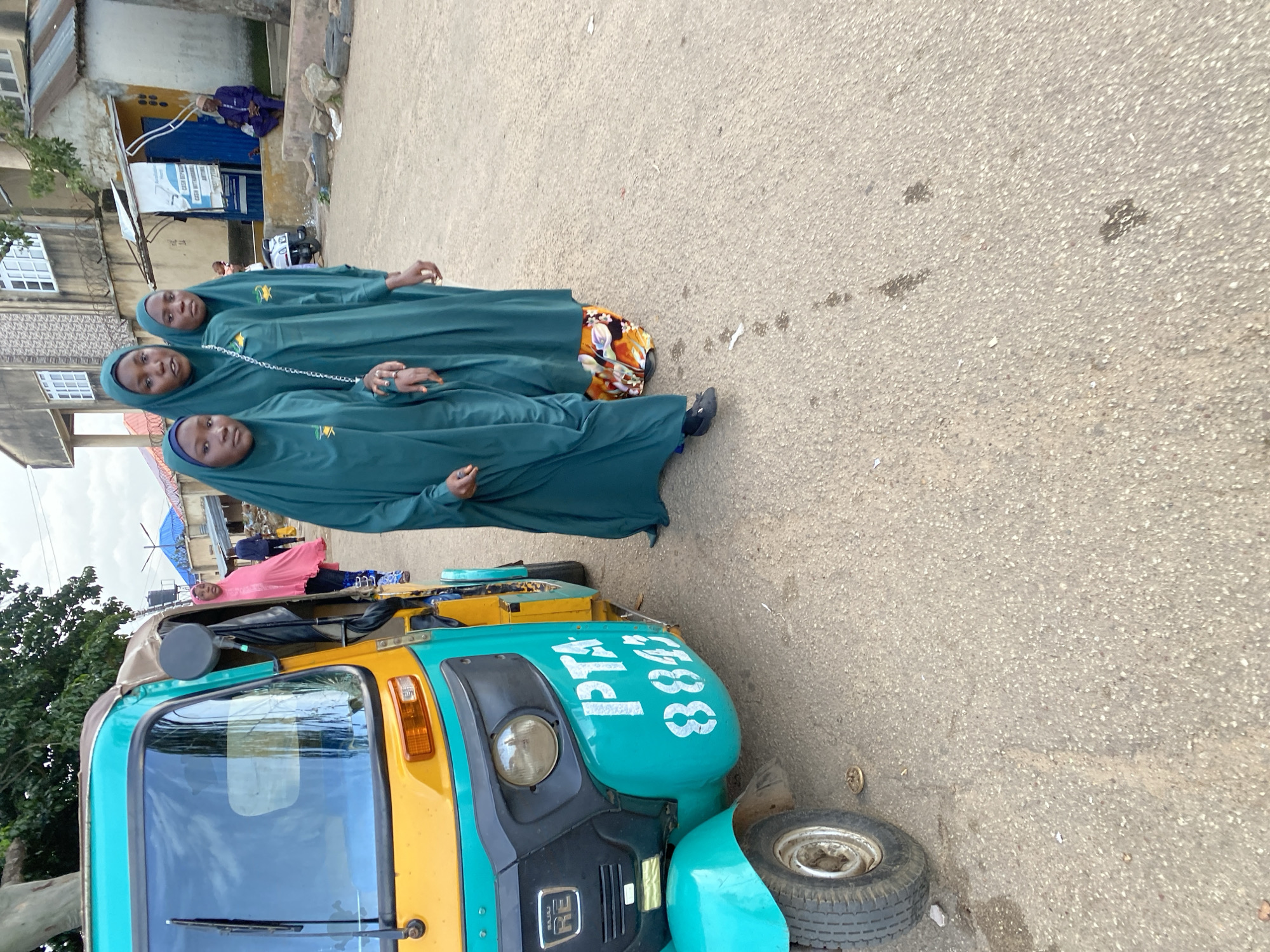Three young women, dressed in dark teal hijabs and long gowns, walk in unison down a paved road in Jos, Nigeria. The woman in the middle has a white chain bag slung over her shoulder. To their left, a brightly colored auto-rickshaw, yellow and teal, is parked. The auto-rickshaw has the markings "PTA" and "8843" in white lettering. In the background, buildings line the street, some appearing to be under construction or in disrepair. A few other individuals are visible in the distance, including a man in a purple traditional outfit sitting on a ledge. The scene appears to be a typical street in a Nigerian urban environment.