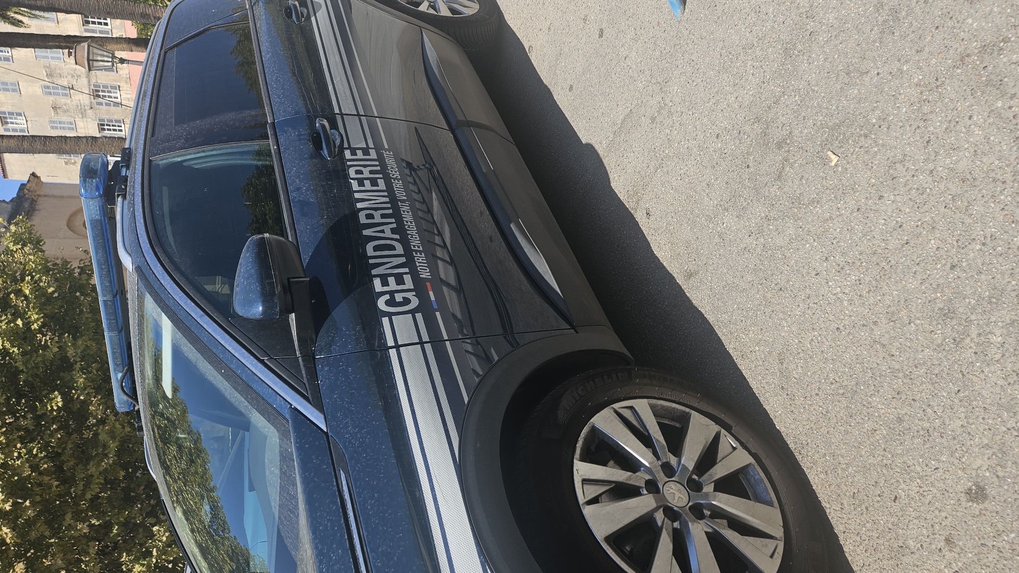 A dark blue Gendarmerie vehicle, likely a SUV, is parked on a light-colored asphalt surface. The side of the vehicle features a reflective stripe pattern and the text "GENDARMERIE" in large white capital letters, with smaller text below that reads "NOTRE ENGAGEMENT, VOTRE SÉCURITÉ." A blue light bar is visible on the roof of the vehicle. In the background, there are trees with green foliage, a building with multiple windows and shutters, and a lamppost. The scene appears to be outdoors in a public area. The overall impression is a static scene, with the vehicle not in motion.