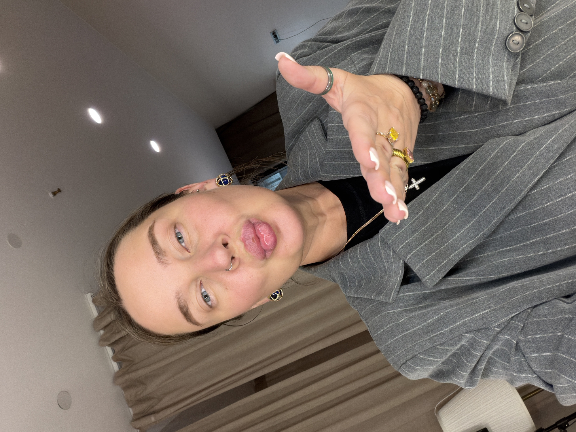 A person with light skin and blue eyes poses for a selfie. They have light brown hair and are wearing a gray pinstripe suit jacket over a black shirt. They have multiple rings on their fingers, including one with a yellow stone, and beaded bracelets on their wrist. A gold necklace with a cross pendant is visible. They have earrings and a nose ring. The background appears to be an interior space with a light-colored ceiling and walls. Based on the available information, the specific location in the city of Khimki, Russia, cannot be determined from the image.