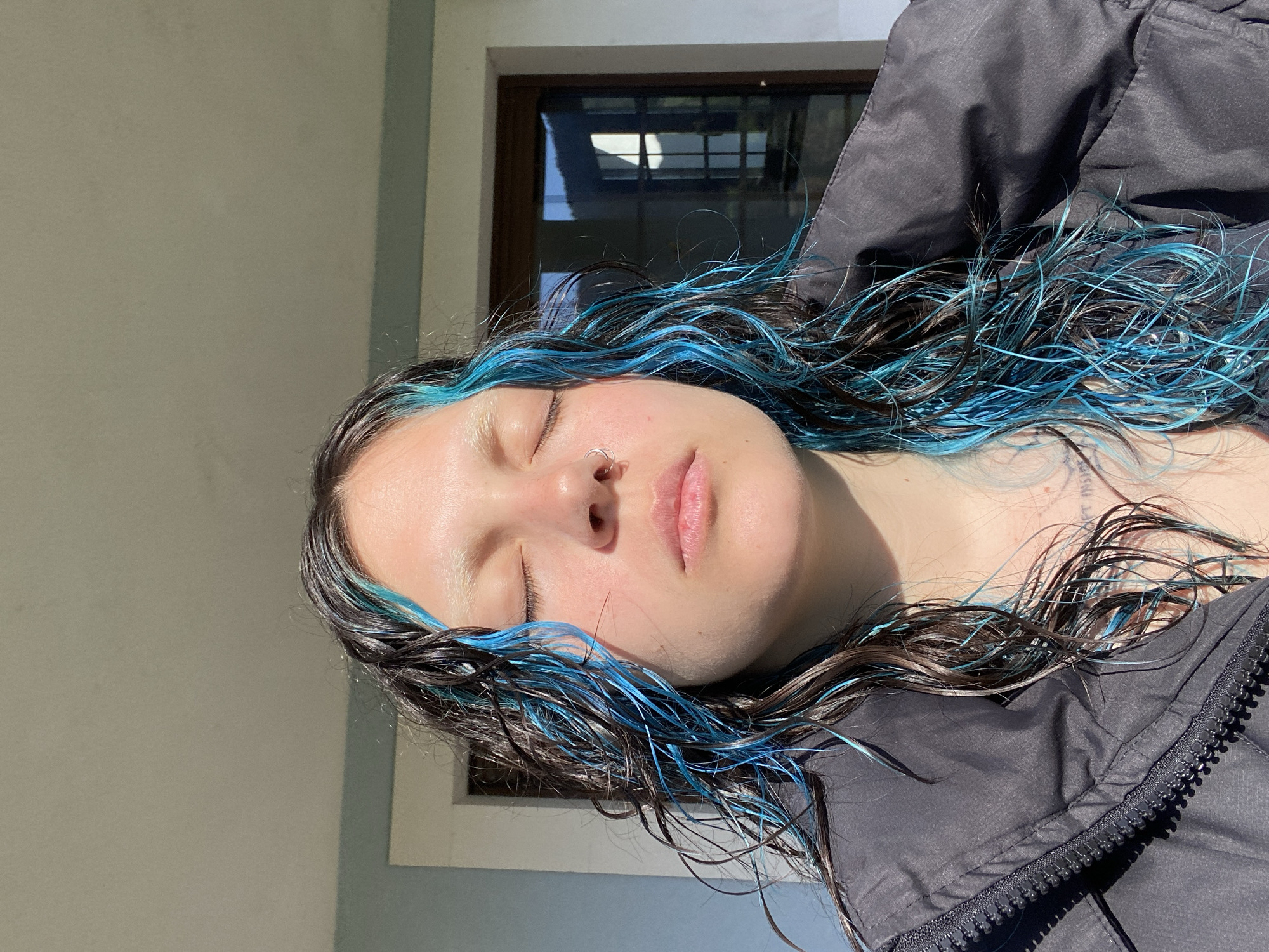 The image is an eye-level, portrait-oriented, medium shot featuring a woman with wet, dark hair streaked with bright blue. She has fair skin and closed eyes, with a silver nose ring visible. She's wearing a dark, possibly black, jacket. The location appears to be outdoors, with a wall visible on the left side of the frame. A window is partially visible in the background. The image shows a strong light source coming from the left, illuminating her face and creating shadows. The presence of the wall and window indicate an urban setting. There is nothing to indicate that the image was taken in Villaviciosa, Spain. Further details would be needed to pinpoint the exact location of the scene.