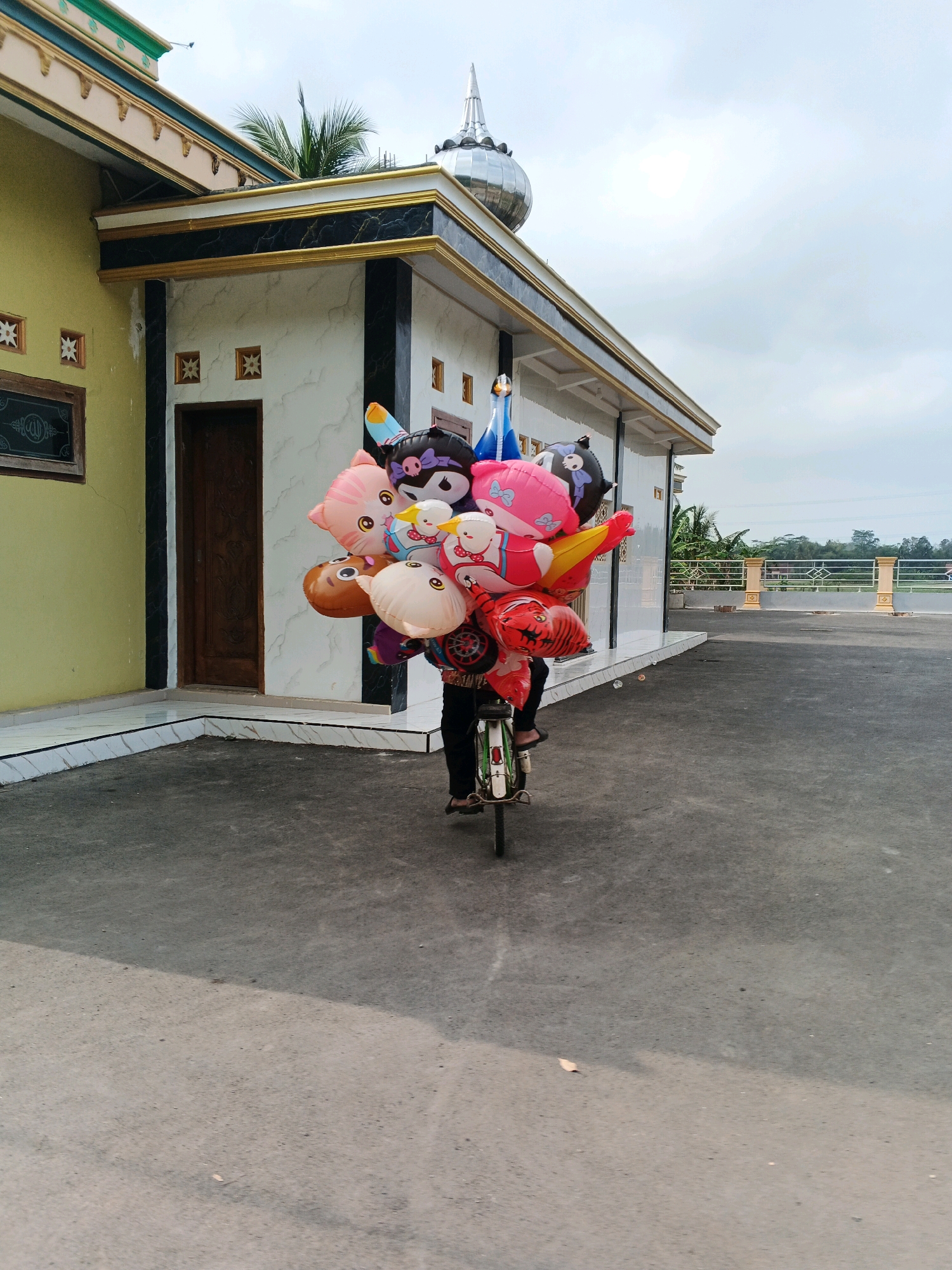 A person is riding a bicycle carrying a large bunch of colorful inflatable balloons. The balloons are shaped like cartoon characters and animals. The person is wearing dark clothing and sandals. The bicycle has a small green frame. The person is on a paved surface in front of a building with a yellow wall and white marbled siding. The building has a decorative roof with a silver dome and gold trim. In the background, there is a metal fence and some greenery. The sky is cloudy.
