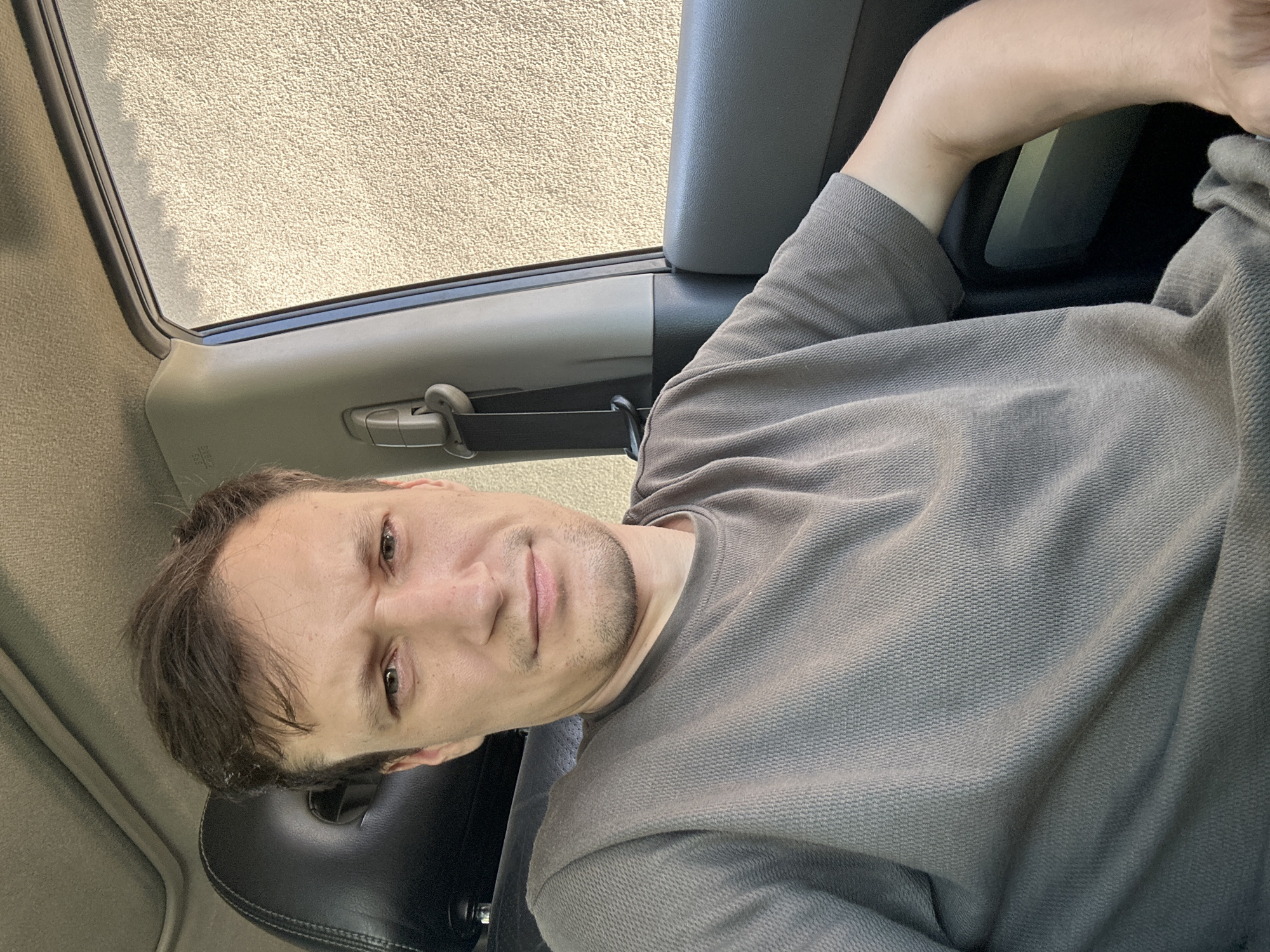 A man is seated in the driver's side of a vehicle, looking directly at the camera with a slightly furrowed brow and a neutral expression. He is wearing a grey, textured t-shirt. The interior of the car is visible, including the headrest of the seat behind him, the car door, a seatbelt, and a portion of the car's ceiling. Through the open car window, a light-colored, textured building wall is visible. The lighting suggests it is daytime. There is no information in the image to suggest this location is in Shatava, Ukraine.