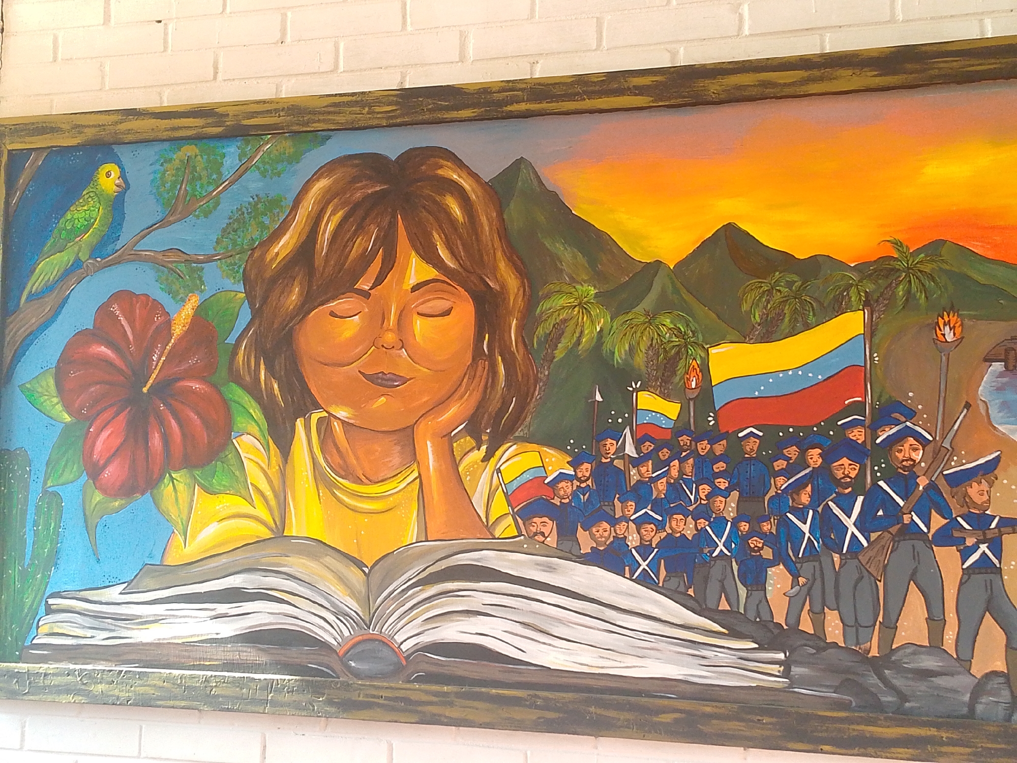 The image is a painting depicting a young woman with brown hair, eyes closed, and a thoughtful expression, her head resting on her hand. She is positioned behind an open book. To her left are a green parrot perched on a branch, a red hibiscus flower, and a cactus. To her right is a scene showing a group of soldiers in blue uniforms with white sashes, carrying weapons and flags, with a backdrop of mountains, palm trees, and a sunset. The flag features the colors yellow, blue, and red, suggesting it could be the Venezuelan flag. The painting is framed and hangs on a white brick wall. The location, Juan Griego, Venezuela, is relevant as the flag and the soldier scene might depict a historical event or a symbol of Venezuelan identity.