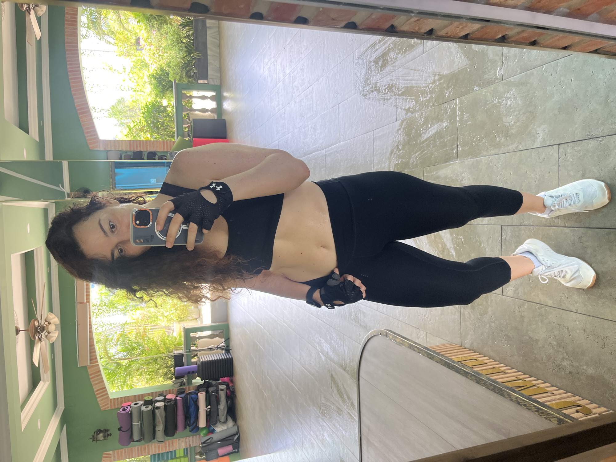 In Ham Thang Ward, Vietnam, a woman is taking a mirror selfie in what appears to be a fitness studio. She is wearing black workout attire, including a sports bra, leggings, and gloves, and white sneakers. The studio has green walls, a ceiling fan, and an arched opening that looks out onto lush greenery and sunlight. Several yoga mats are stacked vertically against a wall in the background, along with other exercise equipment. The floor is tiled and appears damp in places, suggesting recent activity. The woman is holding a smartphone to capture her reflection.