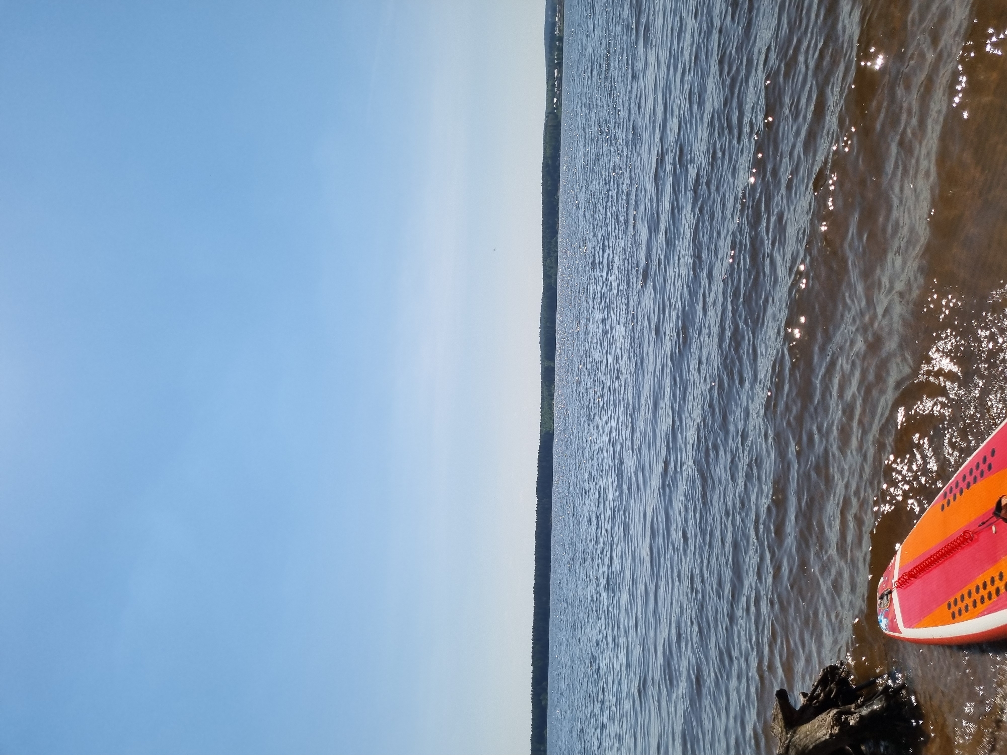 The image shows a body of water, possibly a river or lake, with a shoreline visible in the distance. The water is slightly choppy, with sunlight reflecting off the surface. In the foreground, on the right side of the image, there is a portion of what appears to be a paddleboard, with an orange and red color scheme. The paddleboard is partially in the water, with some of the shoreline visible nearby. The sky is mostly clear with a light blue hue. The city of Chaykovsky, Russia, is not explicitly identifiable in the image.