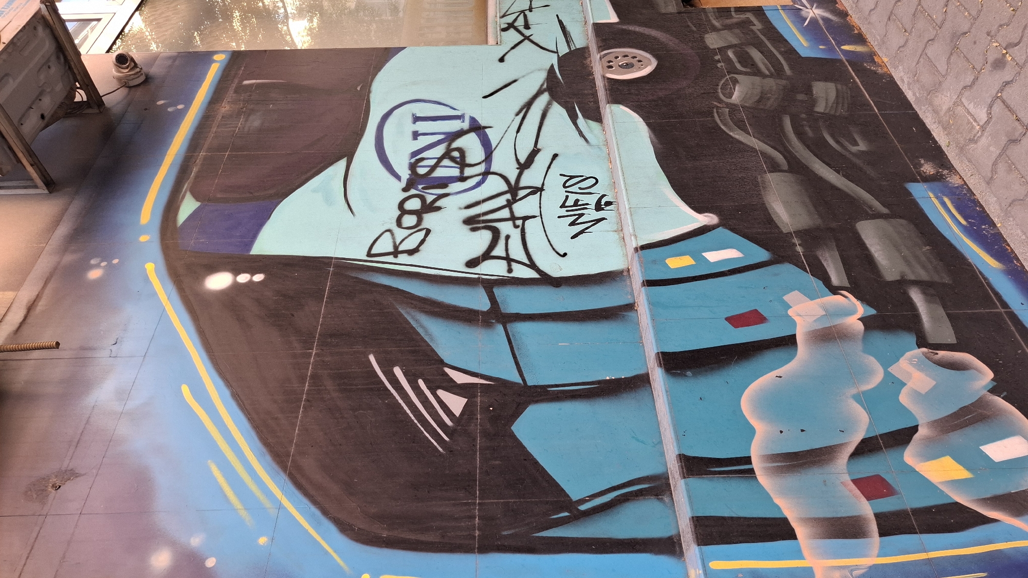 The image shows a graffiti mural on a tiled building facade in Sofia, Bulgaria. The mural features a stylized, abstract design with dominant blue and black colors, accented with yellow lines and white and red shapes. There are several tags and lettering within the graffiti, including what appears to be "BORIS" and "UN", and "WIFI" is clearly legible in white text at the bottom. A section of a car wheel and some mechanical elements are also depicted. The building facade extends upwards, and a security camera and a coiled metal pipe are visible near the top of the image. The ground in front of the mural is paved with light-colored interlocking bricks. The overall impression is of urban street art.