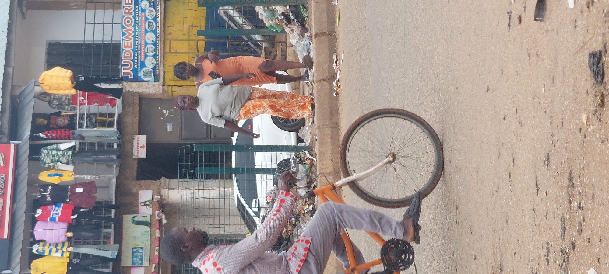 This image depicts an active street scene in the city of Unknown, Unknown.
In the foreground, a man rides an orange bicycle from left to right. He wears a light-colored long-sleeved shirt with red circular patterns and grey trousers, with dark slip-on shoes. The bicycle features a white front fork and dark-colored tires. The road surface is a light-colored asphalt, showing some wear and dark stains.
To the right of the road, a raised concrete curb separates the street from a dirt verge heavily accumulated with various pieces of litter, including plastic bags and papers. A green metal grid fence runs along part of this verge, partially obscuring a white sedan visible in the mid-ground.
On the sidewalk, two women walk from right to left. The woman on the left wears a light grey short-sleeved t-shirt and loose-fitting, orange-and-white patterned trousers. She has reddish-brown braided hair. The second woman, slightly behind and to the right, wears a form-fitting, sleeveless, peach-colored dress and holds a dark mobile phone in her left hand.
The background features commercial buildings. The lower level has a weathered brown facade with a red "Entrance" sign pointing downwards on a white background, and a partially visible sign depicting women's faces, likely for a beauty or cosmetics establishment. The upper level of the building displays numerous items of clothing, including various jackets, shirts (one with a "Supreme" logo), pants, and shorts, hung on rails and displayed on mannequins under a corrugated metal awning. On the right side of the background, a prominent blue sign with white text reads "JUDEMORE GLOBAL INVESTMENT LIMITED," detailing that they are a "DEALER IN ALL KINDS OF CAR SPARE PARTS, BATTERIES, LUBRICANTS, TYRES AND MANY MORE," along with phone numbers. Below this, the sign specifies "OPP. KERAT SUPERMARKET BUKURU MARKET JOS PLATEAU," which provides geographic context for the business advertised. The overall scene is set during daylight hours.