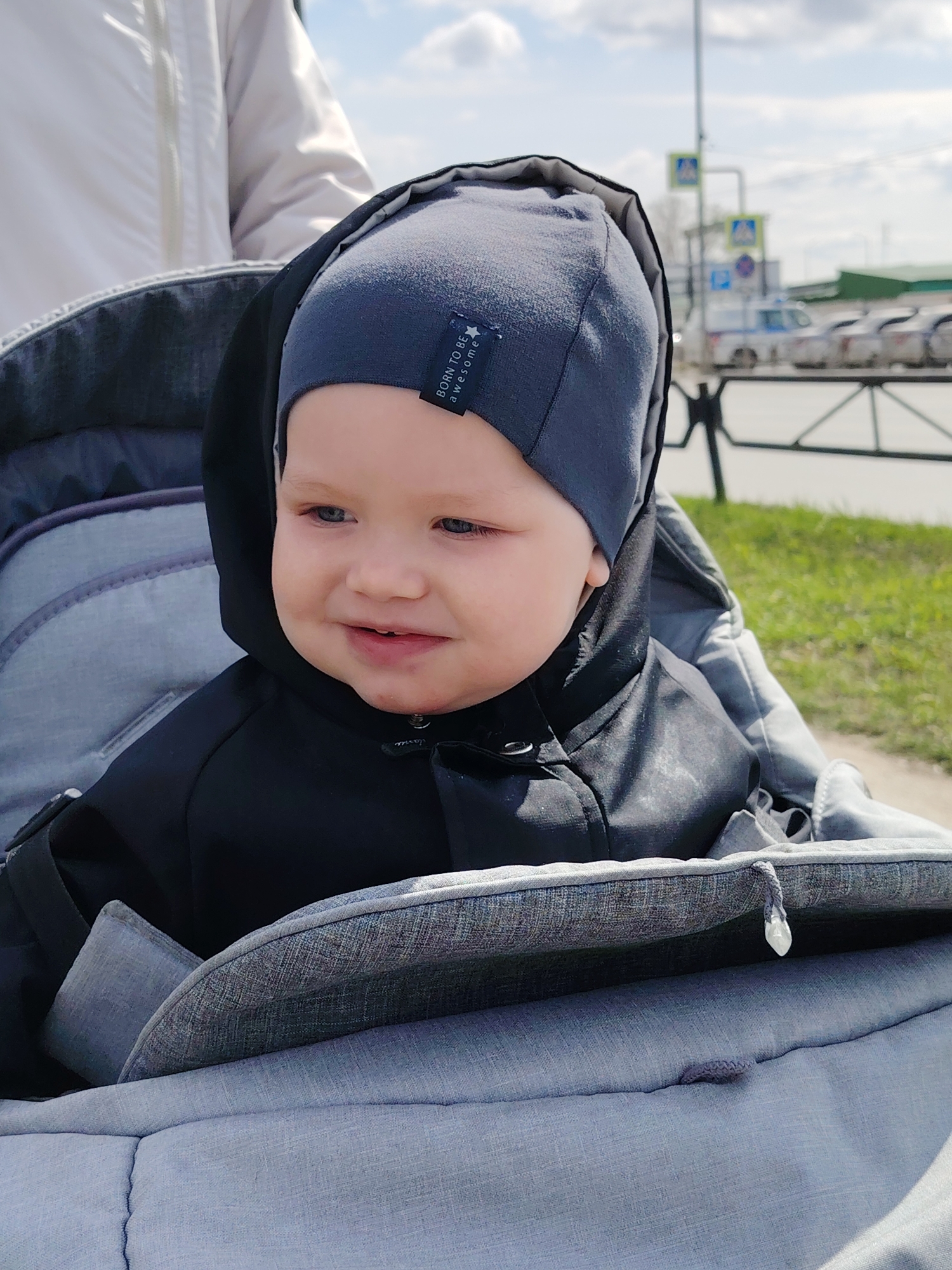 A baby sits in a stroller. The baby is wearing a gray cap and a black hooded jacket. The stroller is gray. Behind the stroller is grass and a road with parked cars. In the background, there is a street sign. The scene is in Chaikovsky, Russia.