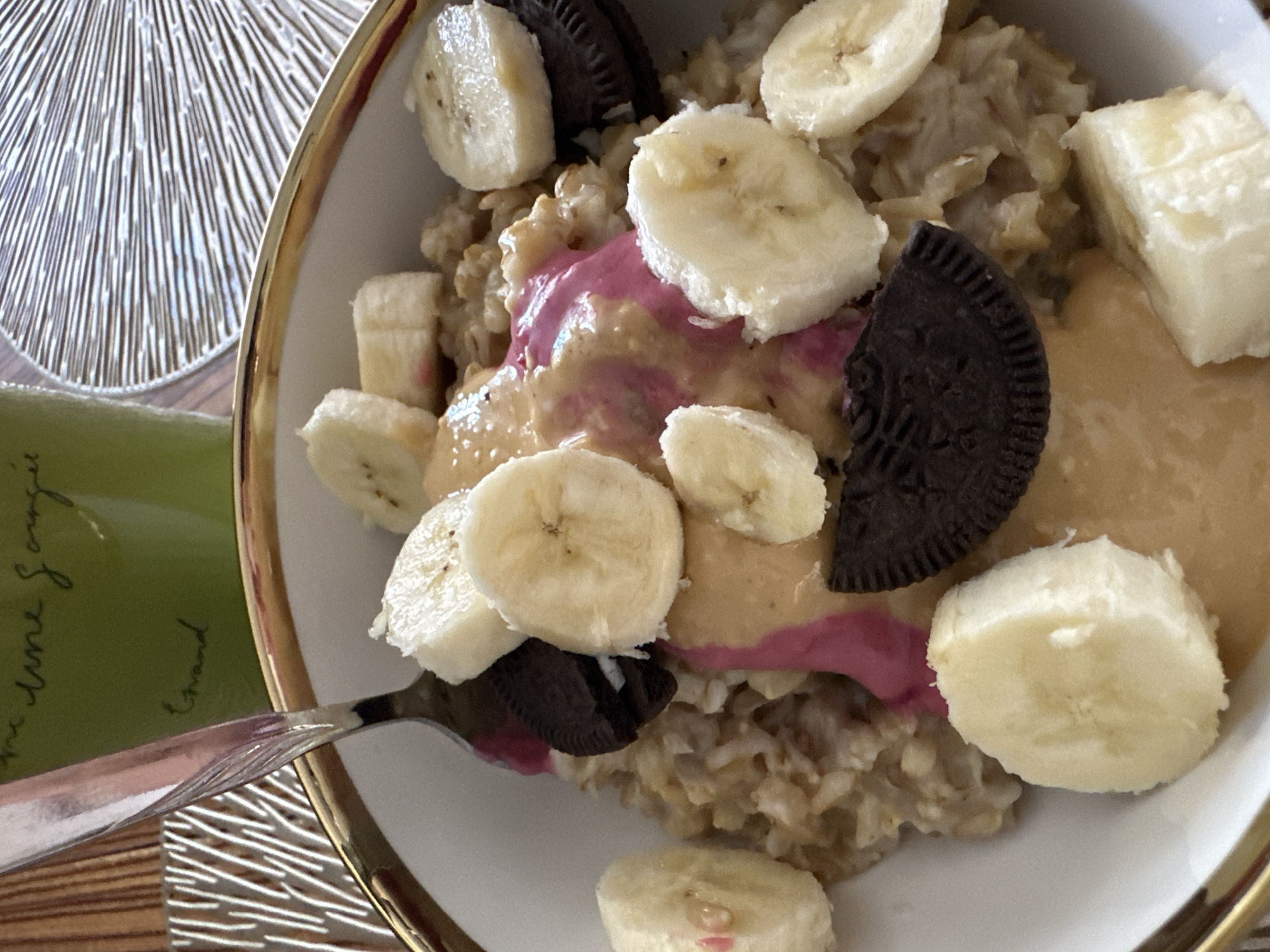 The image shows a bowl of oatmeal topped with sliced bananas, peanut butter, a pink sauce, and Oreo cookies. A fork is partially submerged in the oatmeal. In the background, a green mug with handwriting and a decorative placemat are visible. The scene is indoors, likely in a home or cafe. There is no information in the image to suggest the location is Yuzhnouralsk, Russia.