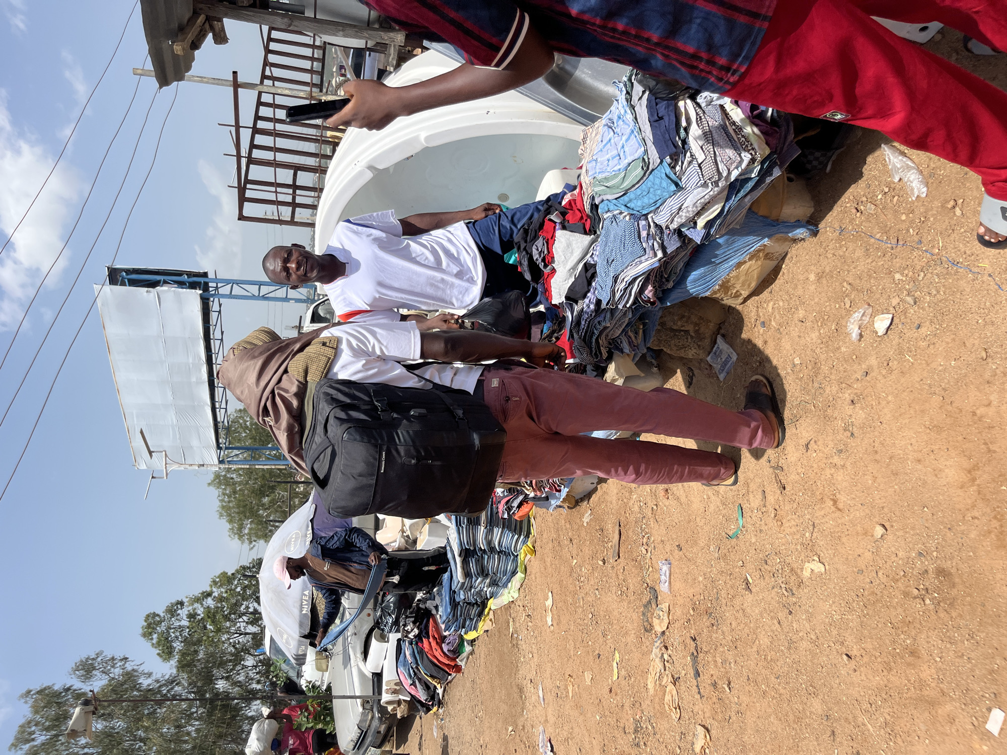 In Gura Top, Nigeria, an outdoor market bustles under a clear, sunny sky. The ground is rough, unpaved earth, scattered with debris. Stacks of folded clothes, primarily shirts and jeans, are displayed on tables and spread out on the ground, forming the primary merchandise.
Several individuals are present. One man, wearing a brown t-shirt and a dark jacket, holds up a piece of blue fabric while standing near parked cars. Another man, in a white t-shirt with red and blue trim, stands facing the camera. A third person, seen from the back, wears a brown head wrap and a black backpack, and maroon pants. A fourth individual, partially visible on the right, wears a red and blue striped shirt and bright red pants, holding up a smartphone as if taking a picture. In the background, a large white billboard stands on a metal frame, and beyond that, trees and a blue sky dominate. A large, white, bathtub-like object is also visible. The scene suggests a typical street-side market environment where goods are sold informally.