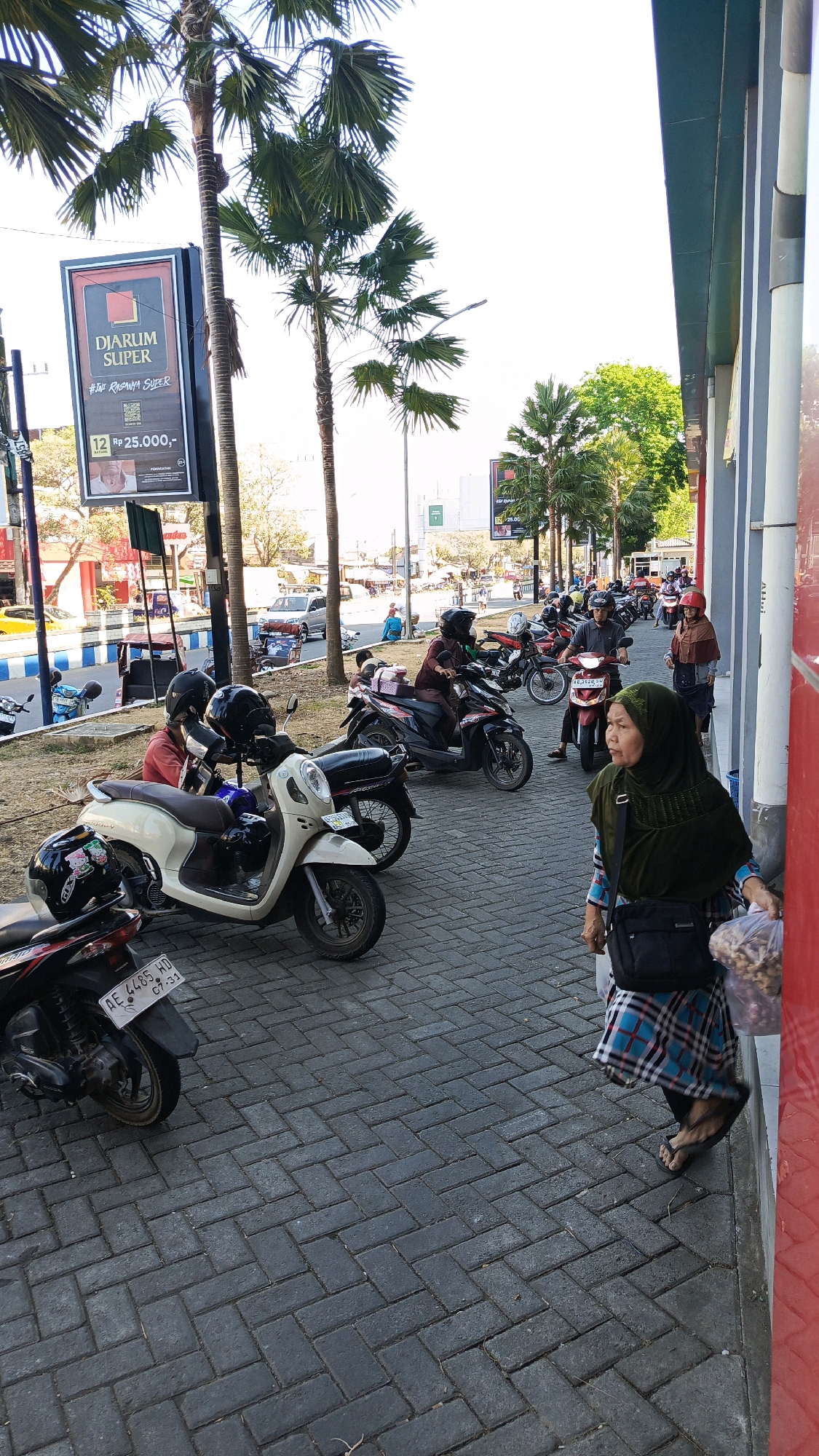 In Ponorogo, Indonesia, a street scene unfolds with several people and parked motorcycles on a brick-paved sidewalk adjacent to a building with a red facade and a gray awning. Palm trees line the sidewalk, and a billboard for "DJARUM SUPER" is visible.
In the foreground, a woman wearing a green hijab and a checkered skirt walks away from the camera, carrying a bag of snacks. To her left, several motorcycles are parked, including a light-colored scooter with black helmets placed on it. Further down the sidewalk, other individuals are seen on or near motorcycles. One person in a maroon outfit rides a motorcycle, and another, wearing a red helmet and brown headscarf, stands near a parked motorcycle. In the background, traffic is visible on the street, with cars and more motorcycles. The sky is bright and overcast.