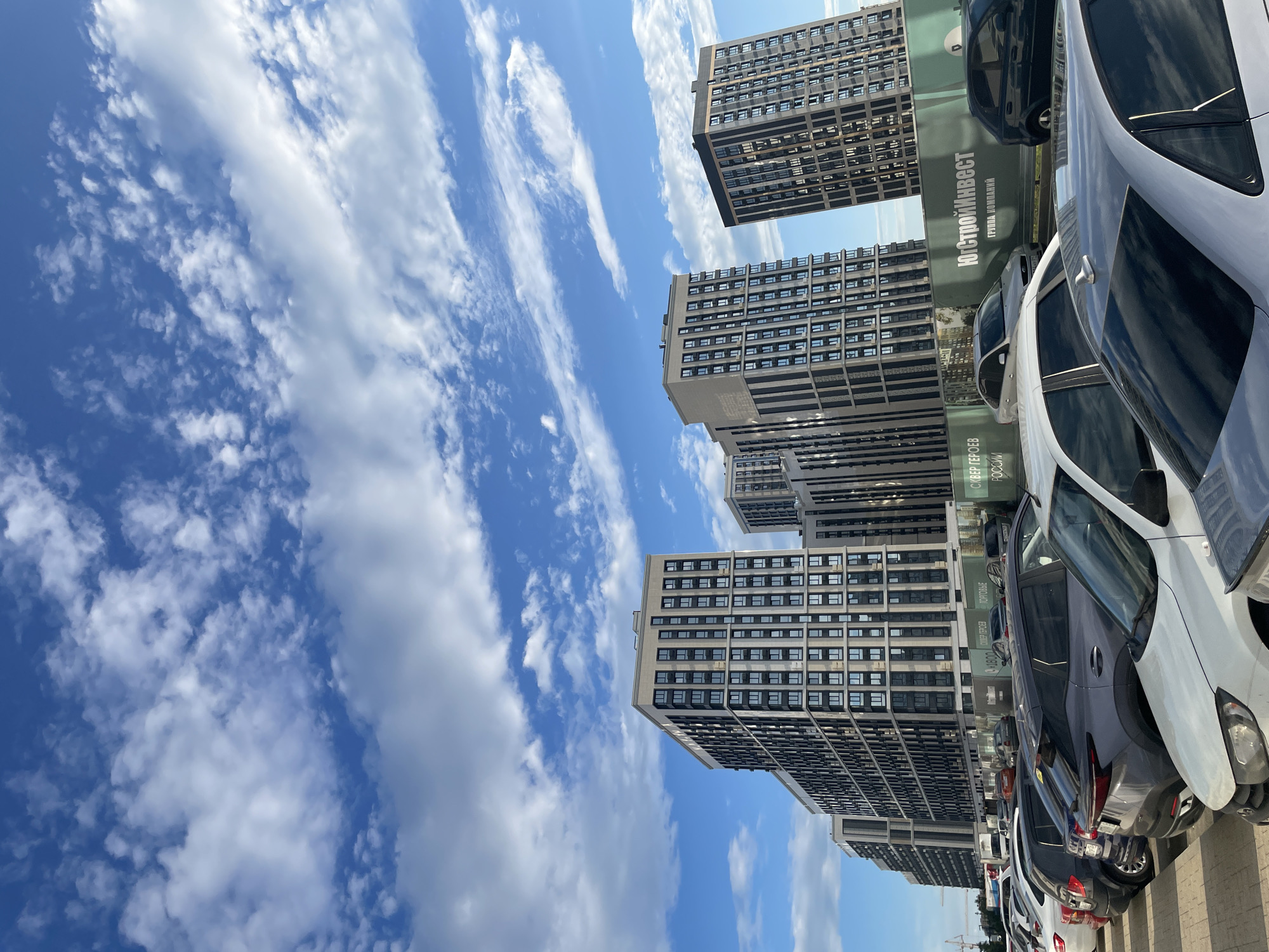 The image depicts a scene in the city of Stavropol, Russia, featuring a modern residential complex and a parking lot filled with vehicles. In the background, several tall, multi-story buildings with a contemporary architectural design dominate the skyline. These buildings are characterized by their repetitive window patterns and a combination of light-colored and dark vertical elements. The sky above is a vibrant blue, adorned with scattered white clouds, suggesting fair weather. In the foreground, a row of cars is parked, indicating a populated urban area. The perspective of the image suggests it was taken from a low vantage point, possibly from within the parking lot or a nearby area. A green fence with white lettering, likely advertising or informational, is visible behind the parked cars, with some Cyrillic text that appears to be "ЮгСтройИнвест" and other related phrases. The overall impression is one of urban development and daily life in a Russian city.