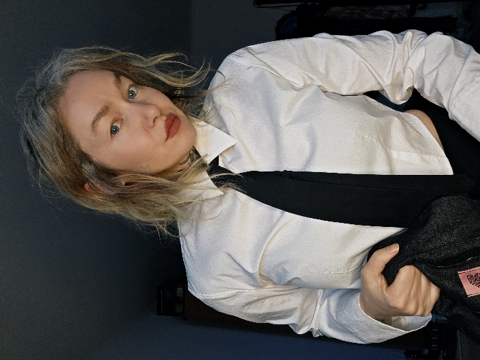 The image displays a person, likely female, from the chest up, positioned at an angle and looking directly at the viewer. The entire image is rotated approximately 45 degrees counter-clockwise.
The person has light-colored, possibly blonde or light brown, wavy hair that falls around their face and shoulders. Their skin tone is fair, and they have clear blue eyes. They are wearing reddish-brown lipstick. A faint tattoo is visible on the left side of their neck, partially obscured by their hair and the collar of their shirt.
They are dressed in a crisp white, long-sleeved button-up shirt with the collar open. A black necktie is worn loosely, hanging down the center of their torso. The shirt appears slightly untucked or loose around the waist. Their left arm is bent, and they are holding a dark, possibly charcoal grey or black, patterned garment, which could be a jacket or sweater. This garment has a small, rectangular pink label with faint, illegible white text on it. Their hand, with manicured fingernails, is visible holding this item.
The background is dark and plain, suggesting an indoor setting. There are some indistinct dark shapes in the upper and lower corners, possibly furniture or objects, but they are not clearly discernible. The lighting appears to be focused on the person, illuminating their face and upper body, creating some shadows on the right side of their face and neck. The overall impression is a posed portrait.