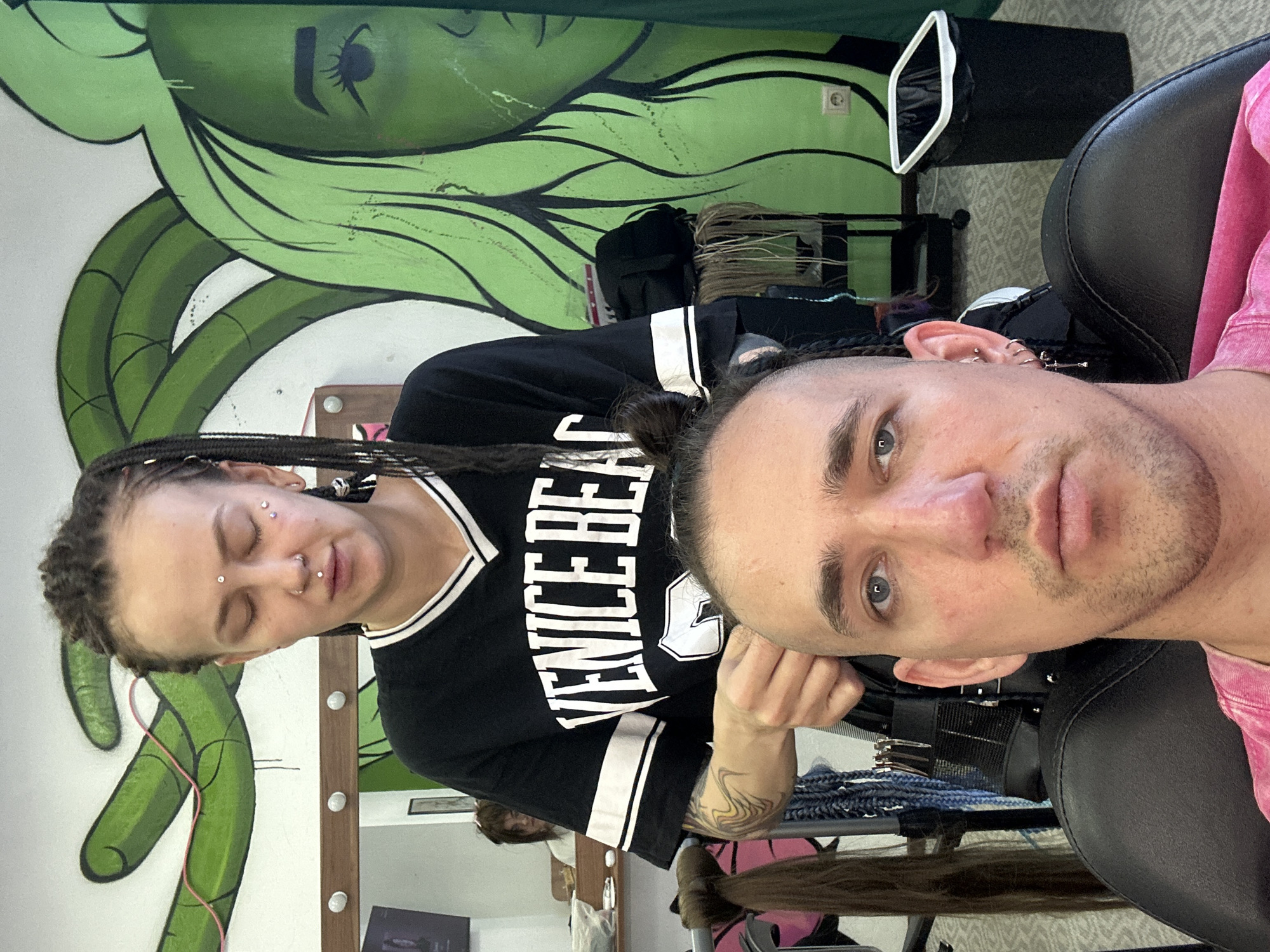 The image shows a close-up of a man with a buzz cut and a woman with braided hair, both facing the camera. The woman is in the process of braiding the man's hair, holding strands of hair in her hands. She is wearing a black t-shirt with white stripes on the sleeves and the text "VENICE BEACH" printed on the front. She has piercings on her nose, lips, and forehead, and various other facial piercings. The man is wearing a pink shirt and has blue eyes. He also has several earrings. Behind them, a mural of a green face with flowing hair is painted on the wall. In the background, a mirror reflects another person, and hair extensions and styling tools are visible on shelves and a trolley. A black trash can with a white rim stands to the right of the frame. The overall environment appears to be a hair salon. No specific location in Saratov, Russia is indicated in the image.