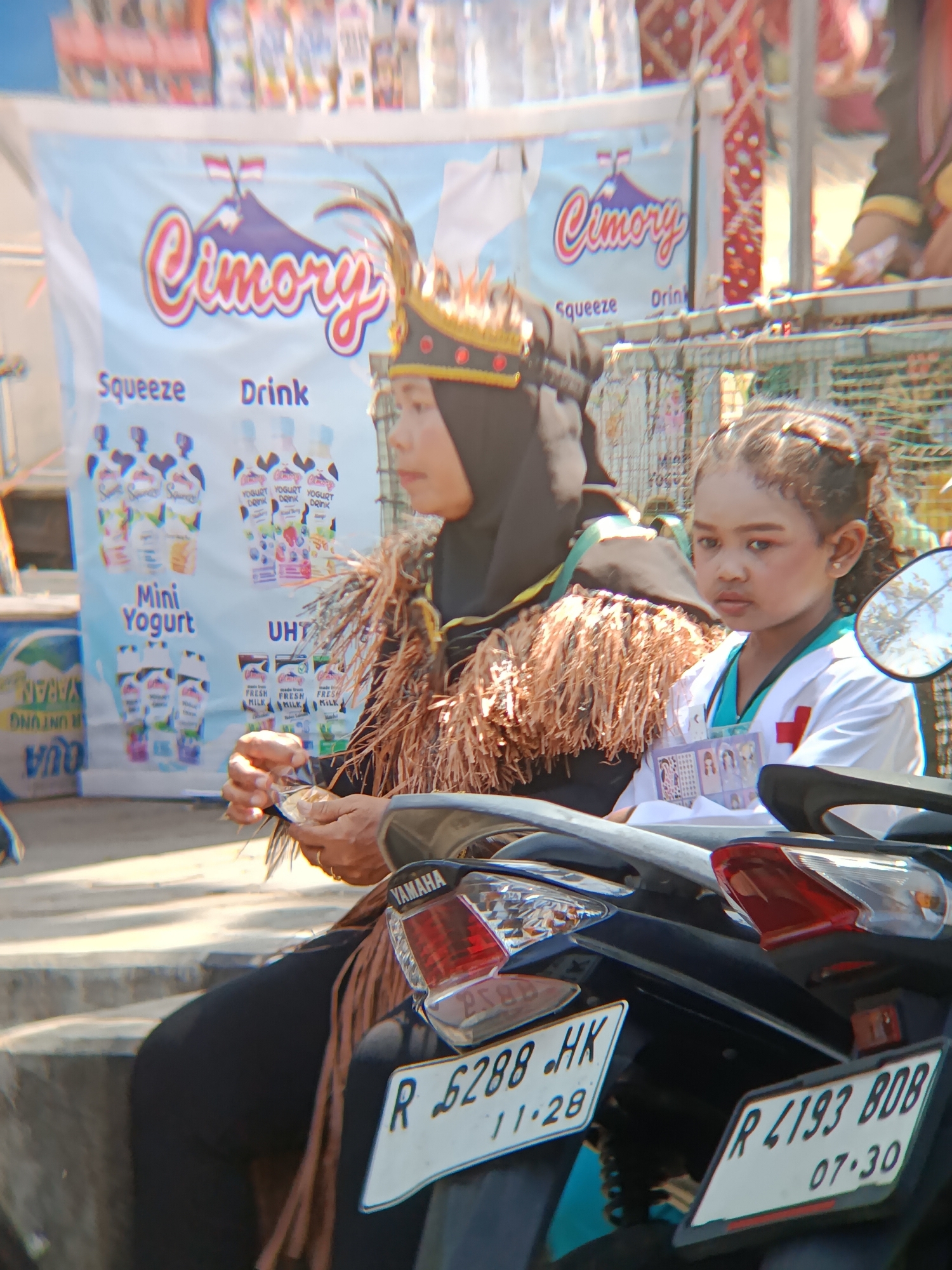 In the village of Desa Kebon Manis, Kecamatan Cilacap Utara, Indonesia, two individuals are captured on a motorcycle. In the foreground, a person, potentially an adult, is seated on the motorcycle, wearing a black headscarf and an elaborate headdress adorned with feathers. They are dressed in a brown, fringed garment over black clothing and are holding a small, silver packet. Beside them, a young child is also seated on the motorcycle, dressed in a white doctor's coat with a red cross emblem. The child has makeup applied, with noticeable blush and eyeshadow. Behind them, a banner displaying the "Cimory" logo and product images for yogurt and milk is visible. The background suggests an outdoor setting, possibly a market or event, with other stalls or displays partially visible. The license plate on the motorcycle indicates it is from Cilacap.