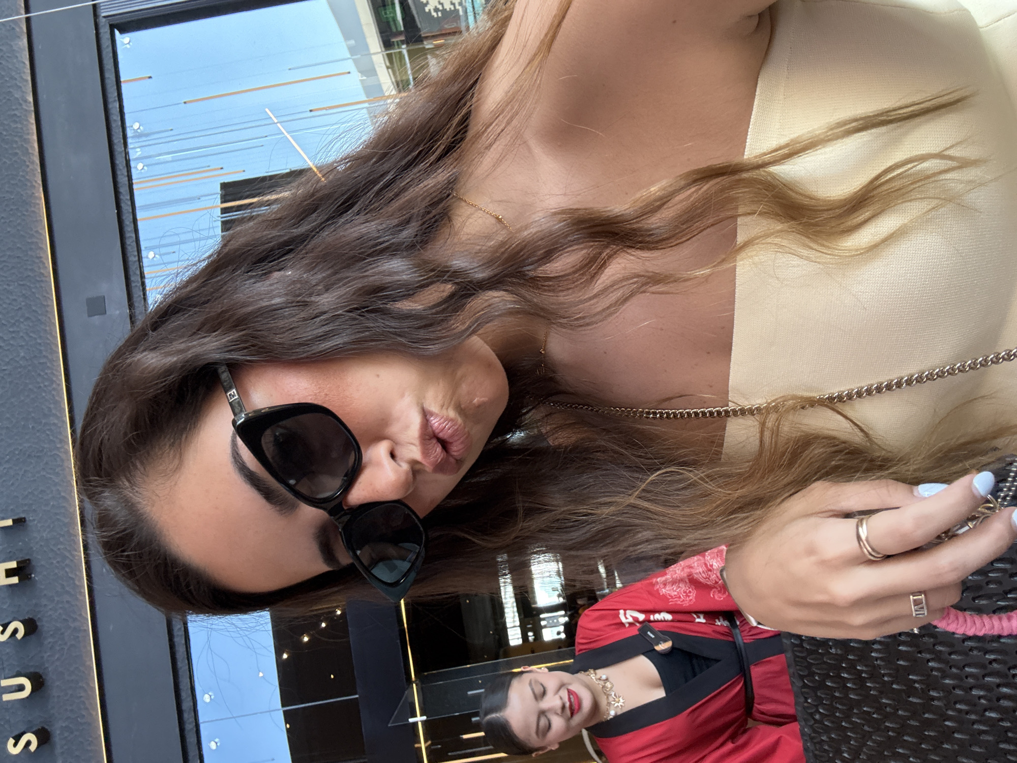 A woman wearing sunglasses and a light-colored top is seated at a sushi restaurant, as suggested by the vertical "SUSHI" sign visible in the background. She is looking at the camera with her lips pursed. Another woman, wearing a red and black outfit with gold jewelry, is visible in the background. The photograph appears to have been taken outside of the restaurant and the buildings are reflected in the glass, suggesting it is located in San Bernardino Tlaxcalancingo, Mexico. The woman is holding a dark-colored bag or object in her hand, and her fingers are adorned with rings, she also has light blue nail polish.