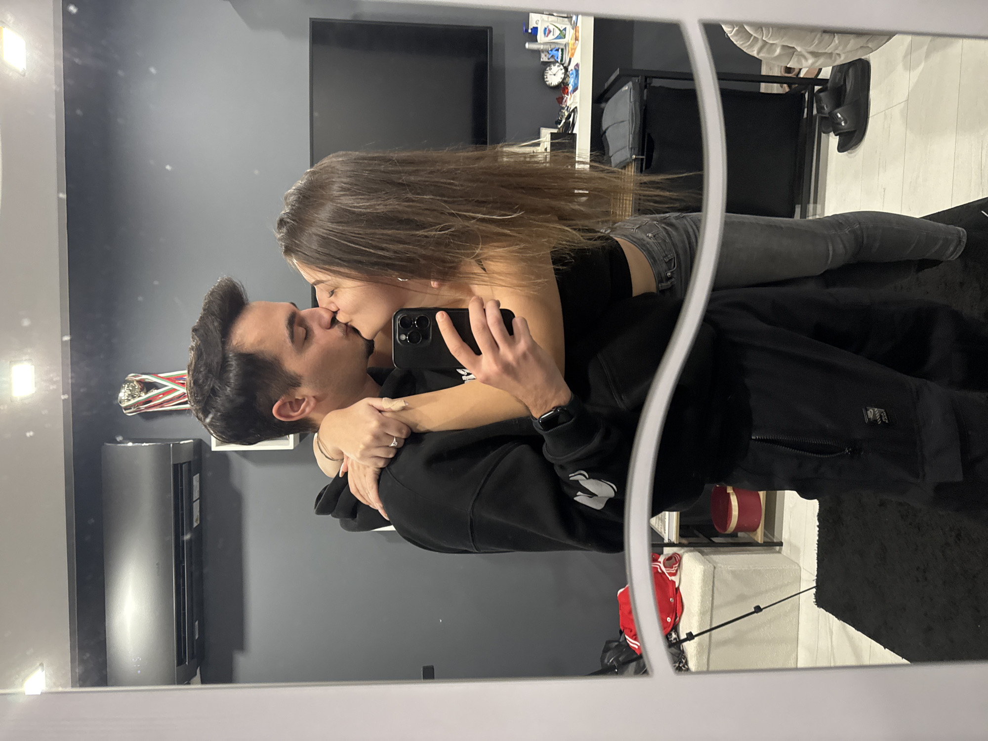 A couple is kissing while one of them is holding a phone, presumably taking a picture of themselves in a mirror. The woman has long brown hair, a black top, and grey jeans. The man is wearing a black hoodie and has dark hair. The environment appears to be a room with grey walls. A television is visible on the wall, along with an air conditioner and some shelving. Based on the style of the interior, the scene is likely taking place in a residential apartment. The city and location are not discernible from the image.
