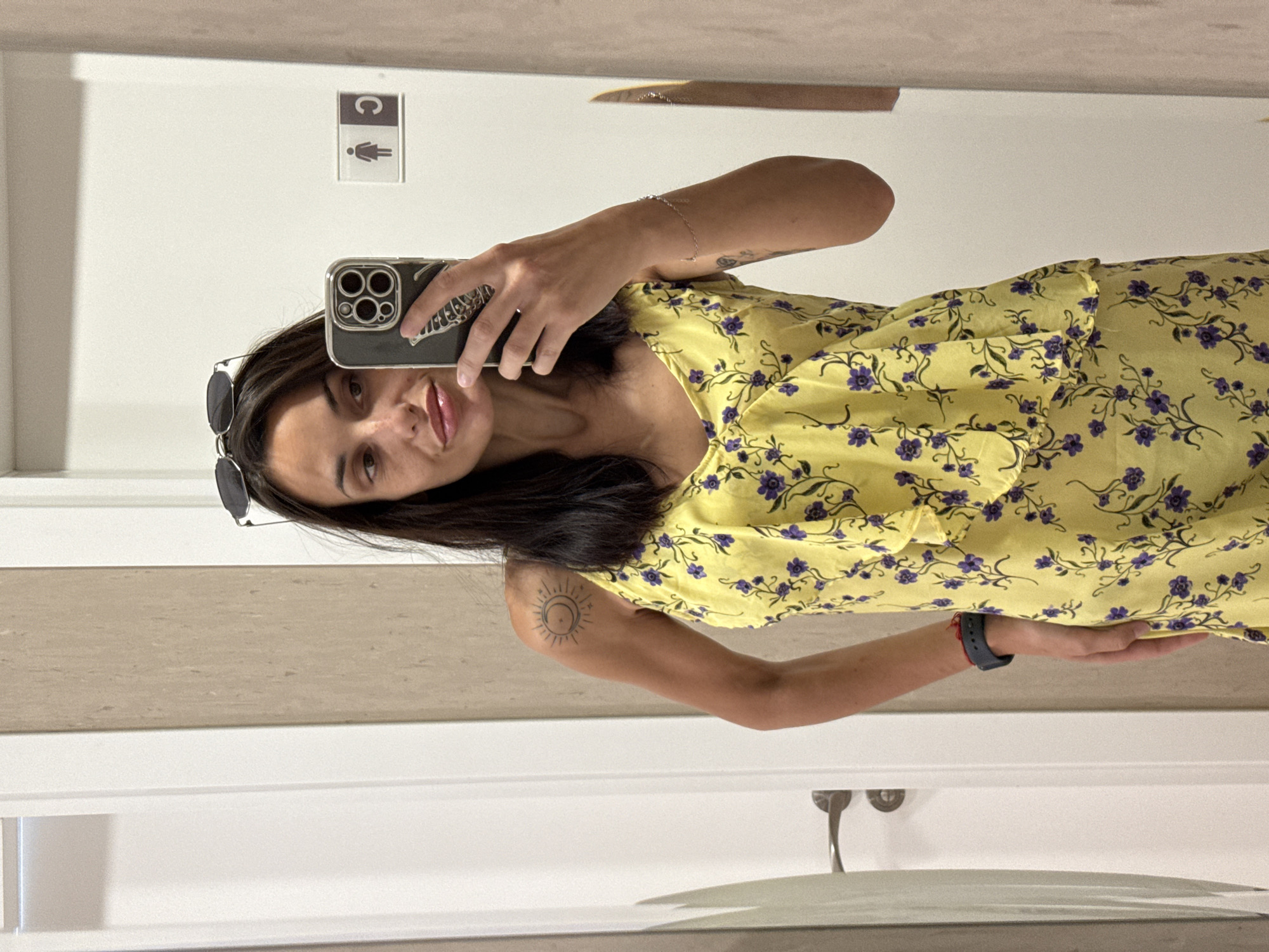 A woman with dark hair and sunglasses perched on her head is taking a mirror selfie in a fitting room. She is wearing a yellow floral print dress. She has a sun tattoo on her left shoulder. There is a sign on the wall indicating a restroom. The fitting room appears to be in a retail establishment in Pleven, Bulgaria.