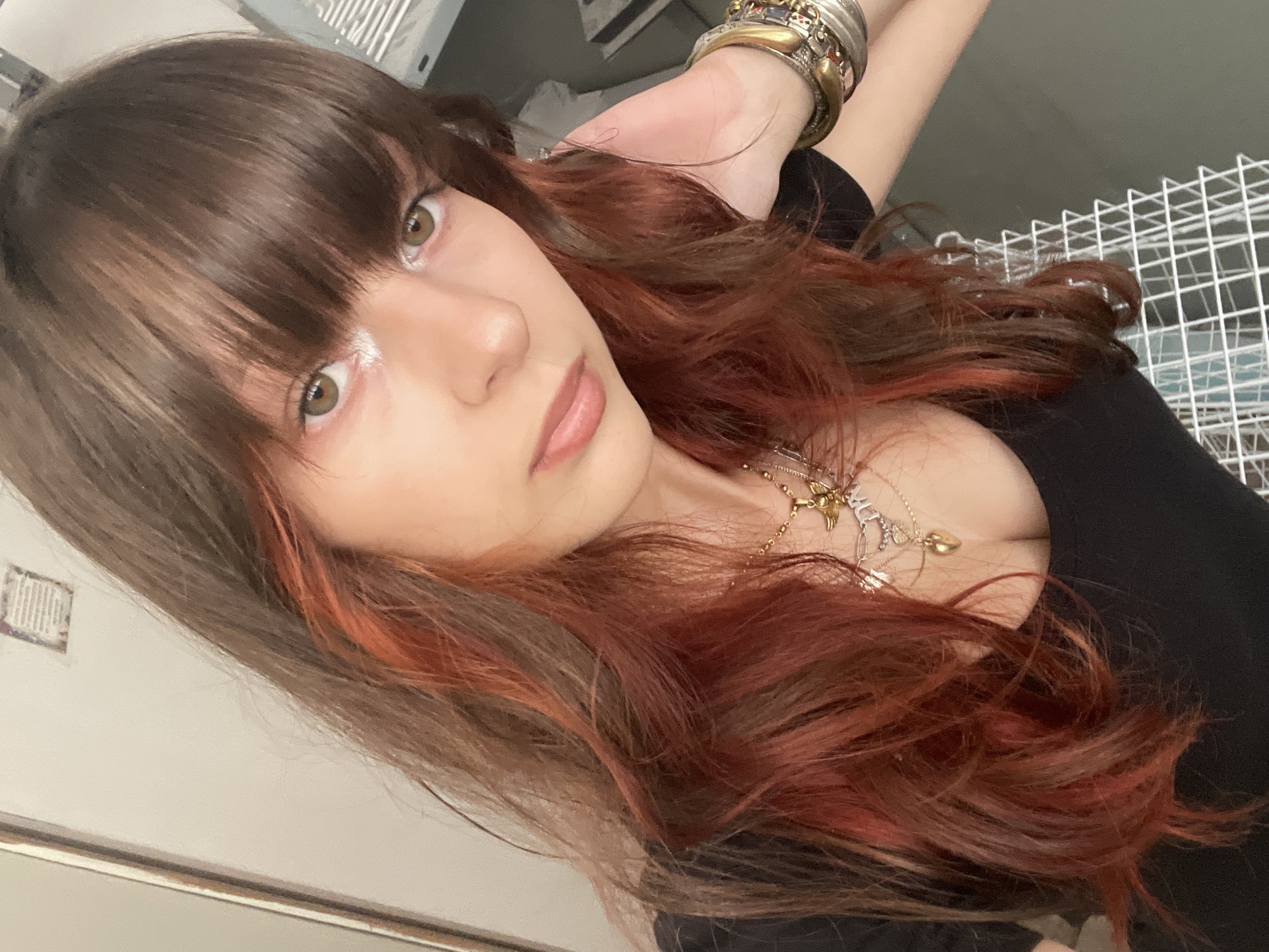A medium close-up, angled selfie depicts a young woman with fair skin and brown hair with auburn highlights. She has straight bangs and is looking at the camera. Her eyes are light brown. She appears to be wearing white makeup or glitter on her forehead. She is wearing a black top. Several necklaces hang around her neck. Her left wrist is adorned with multiple bracelets. The background shows parts of a room with white shelves and a wall with a framed picture. Based on the request for a city location, the photograph appears to have been taken indoors in a private residence or commercial location. The information available does not corroborate a location in Raanana, Israel.