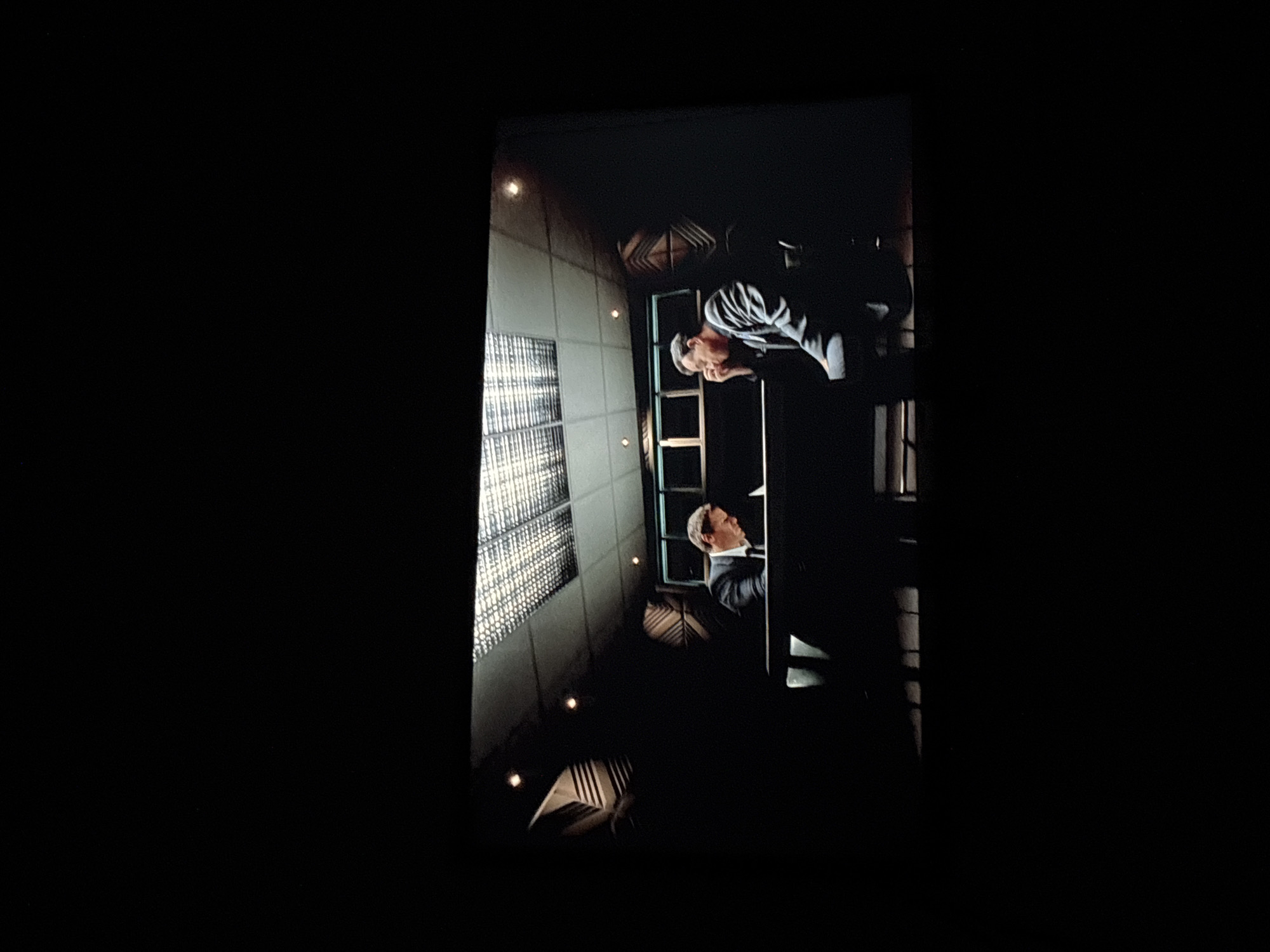 The image shows a scene from a film, possibly a dramatic or suspenseful one. Two men are visible, seemingly in an office or institutional setting. One man is wearing a dark suit and a light shirt. The other man, positioned closer to the window, is wearing a dark striped jacket. The background features a wall with a large window, likely made of frosted glass, providing natural light. There is an artificial light source also. It's difficult to ascertain the specific location in Buenos Aires without more information. Additional details like architectural style, signage, or any elements specific to the city are missing.