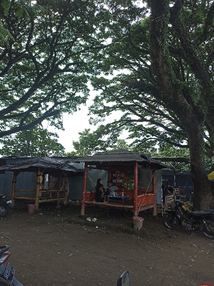This outdoor image from Kepung, Indonesia, captures a rustic gathering area nestled under a dense canopy of large trees. The scene is dominated by several simple, open-sided huts or shelters, constructed from bamboo and wood, with roofs made of corrugated iron or tarps. The ground beneath is unpaved dirt.
In the central, red-painted bamboo hut, an adult woman with dark hair sits with two children, a boy and a younger child or baby, at a low table. They appear to be relaxing, possibly enjoying food or drinks. On the back wall of this hut, a sign is prominently displayed with the text "RM RUMAH MAKAN" (Restaurant) and "MAU DI MARI" (likely "Want to come here" or "Come here"), along with some colourful, food-related graphics. To the left, an empty, natural bamboo hut stands with similar construction. Another individual is partially visible sitting in a more basic shelter to the right, behind the central hut.
The upper portion of the image is filled with the sprawling, leafy branches of large trees, creating a natural, shaded ceiling. The bright, overcast sky indicates it is daytime, providing soft, diffused light across the scene. Parts of two motorcycles are visible, one in the lower-left corner and another parked to the right, suggesting it might be a popular stop-off point. A pair of white flip-flops can be seen on the ground near the central hut. The overall atmosphere is casual and tranquil, characteristic of a local roadside eatery or resting spot.