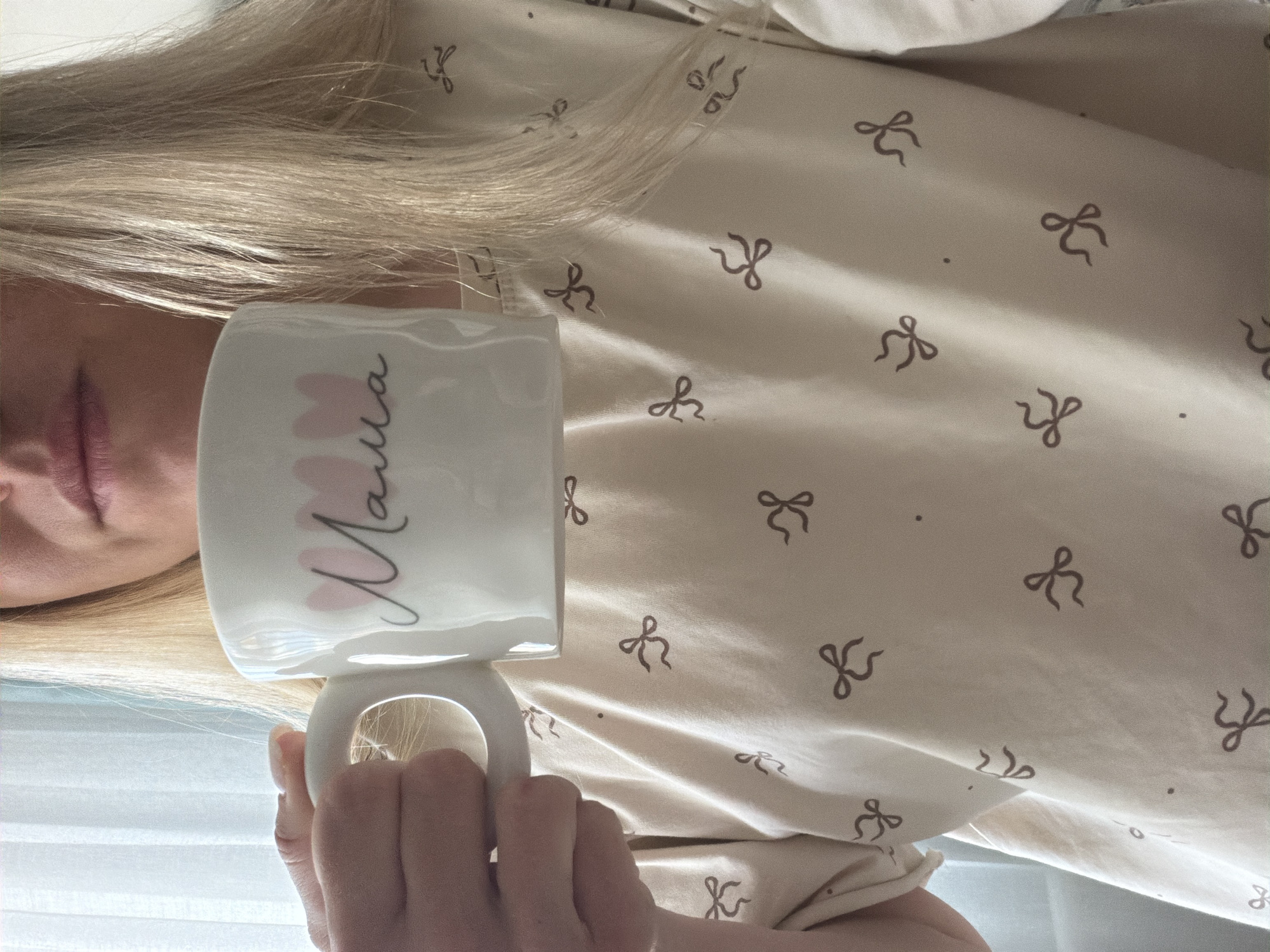 A person with blonde hair is holding a white mug with the word "Mama" written in Russian in pink and black letters. The person is wearing a light-colored shirt with a pattern of small brown bows and dots. The scene appears to be indoors, possibly near a window with blinds. There is no information to suggest that the location is in Votkinsk, Russia.