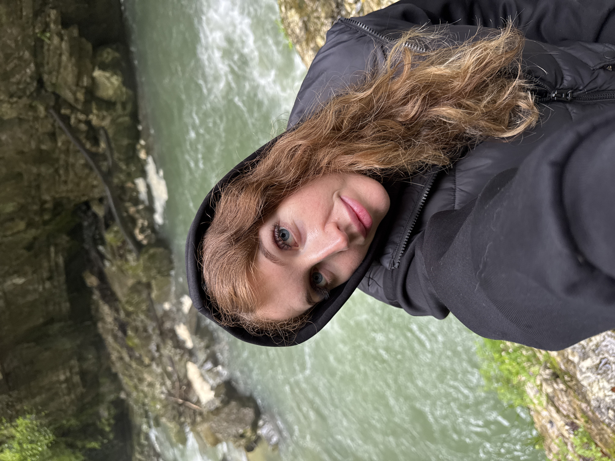 A person with light skin and long, light brown, wavy hair, wearing a black hooded jacket, takes a selfie in Tsabal, Abkhazia. The background shows a river or creek flowing through a rocky, moss-covered canyon.