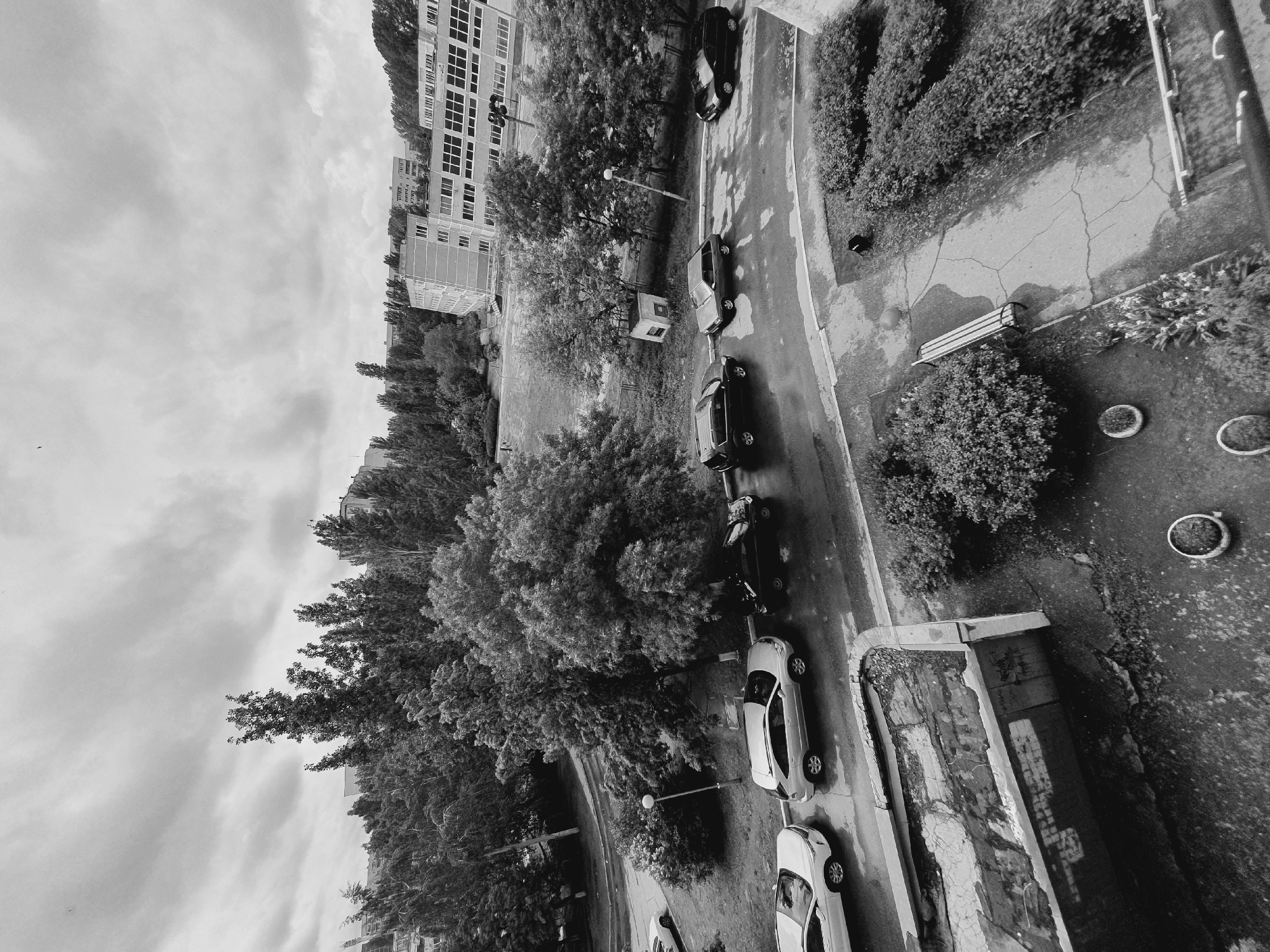 A black and white, high-angle photograph depicts a residential street scene in Ulyanovsk, Russia. The perspective is rotated ninety degrees clockwise. The street runs vertically through the center of the frame and is lined with parked cars on both sides. A row of buildings is visible along the left edge of the frame, partially obscured by trees. Along the right side of the frame, a sidewalk area with benches and landscaping is present. The sky is overcast.