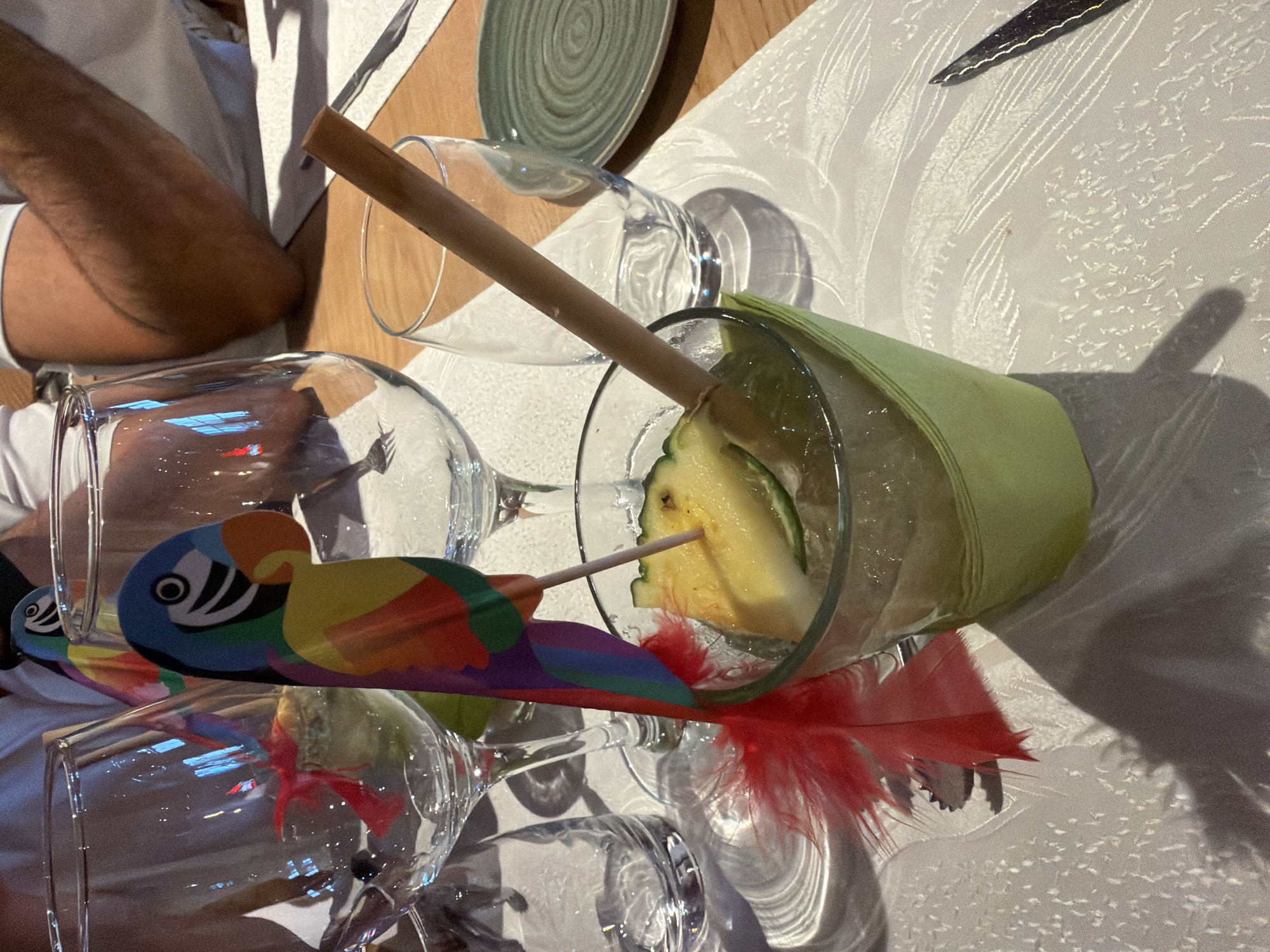 The image shows a close-up view of a table setting in a restaurant. In the foreground, a glass contains a drink with ice and a slice of pineapple and lime, garnished with a colorful parrot-shaped toothpick with a red feather. A wooden straw is also in the glass. The glass is wrapped in a light green napkin. Behind the drink, there are several empty wine glasses and a small plate. The table is covered with a white tablecloth with a textured pattern. In the background, parts of people's arms and torsos are visible, suggesting they are seated at the table. The lighting suggests an indoor setting, likely during the day or evening. The general ambiance appears to be festive, possibly due to the colorful garnish on the drink. The photograph was taken at the restaurant "L'Express" in Fentange, Luxembourg.