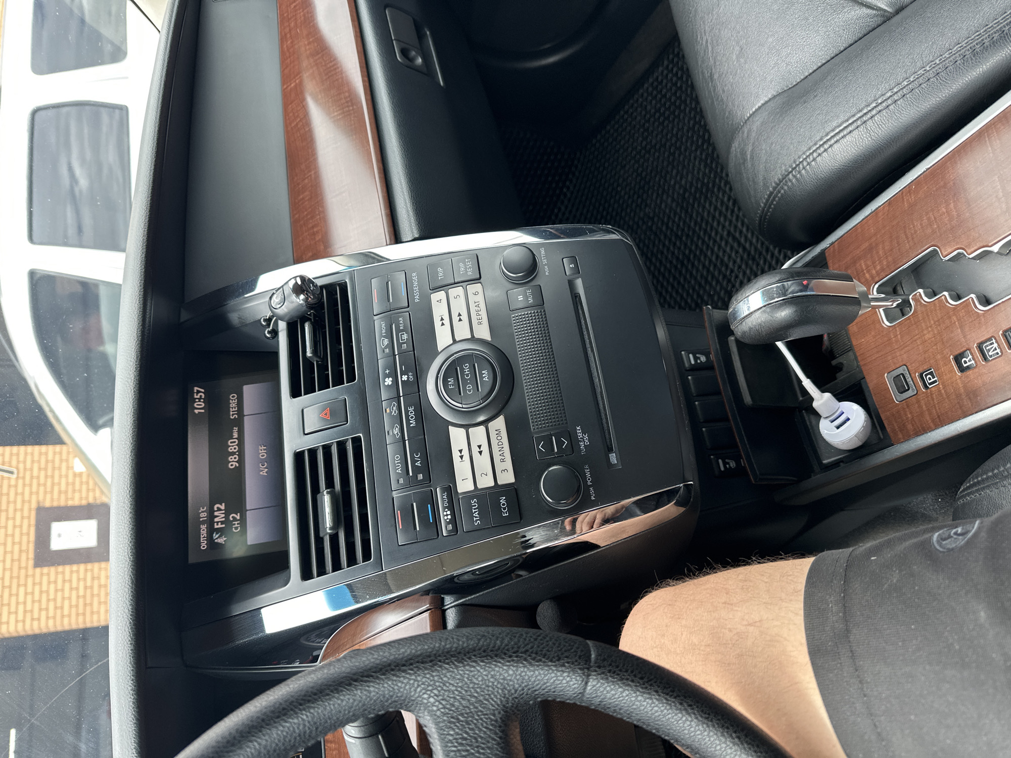 The image shows the interior of a car. The focus is on the dashboard and center console, with a view of the steering wheel and a portion of the driver's leg. A digital display shows the time as 10:57, outside temperature as 18°C, and radio station information. Buttons and controls for the car's audio system and climate control are visible, including options for FM, CD, AM, A/C, and mode settings. A gear shifter is also visible, with markings for Park (P), Reverse (R), and other gears. A USB charger is plugged into a port. The interior features wood trim accents. The scene appears to be set during the daytime, likely under natural light. The car's interior is clean and orderly. The visible text on the dashboard includes various labels for the car's functions and settings. No people are clearly depicted, though a leg and knee are visible, suggesting a driver or passenger. The overall context is a person inside a parked or stopped car.
