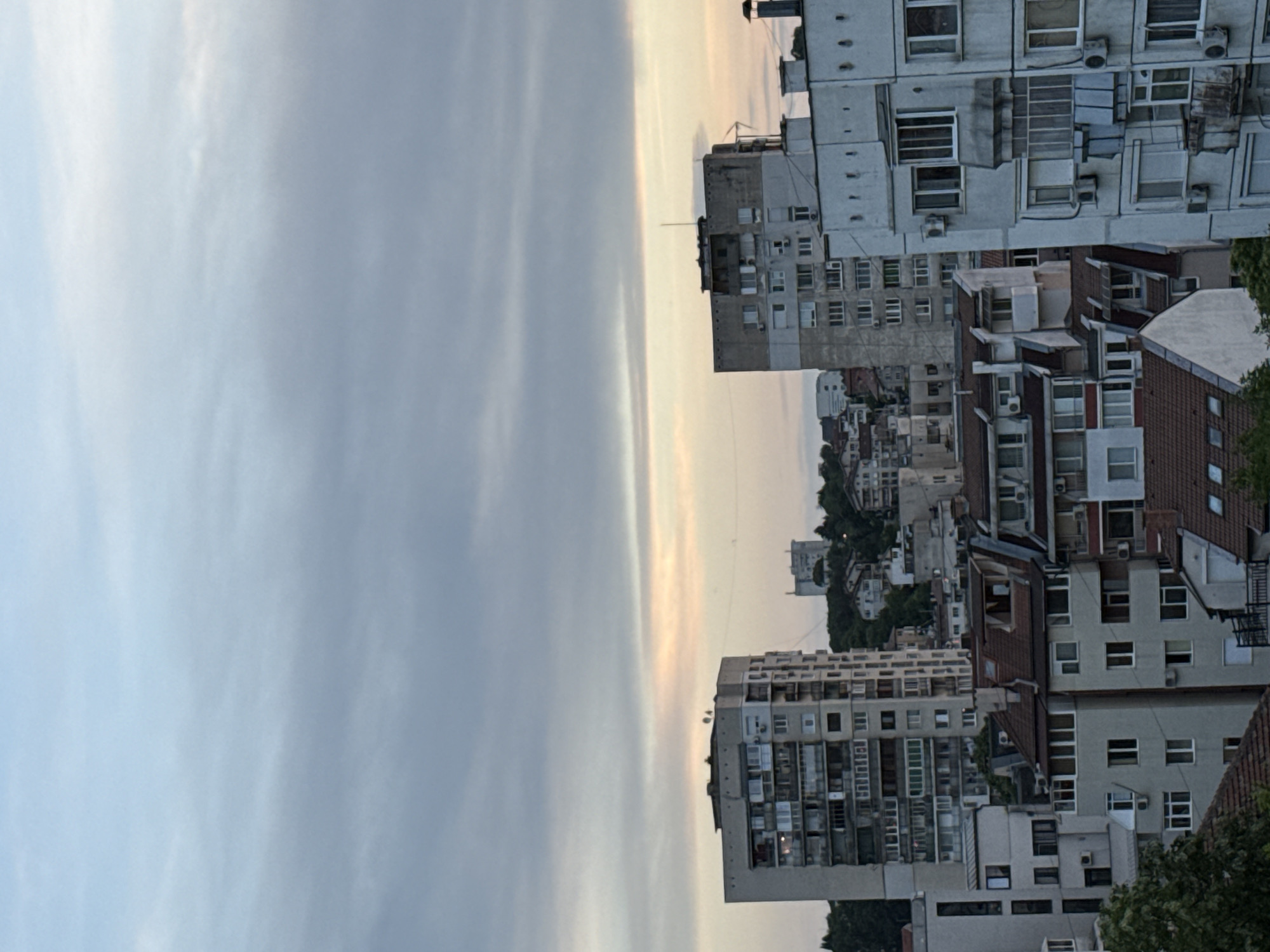 The image presents a cityscape viewed vertically, with a broad expanse of sky dominating the left side. The sky displays varying shades of gray, suggesting an overcast or twilight condition. The city, situated on the right, is characterized by a mix of residential buildings of varying heights and architectural styles.
Several mid-rise apartment buildings are visible, featuring repeating window patterns and rooftop structures. The buildings are constructed with materials and designs typical of urban residential areas. No people are discernible in the image.
While the image captures the general scene, specific details about the city of Pleven, Bulgaria, are not clearly distinguishable to precisely pinpoint the location.
