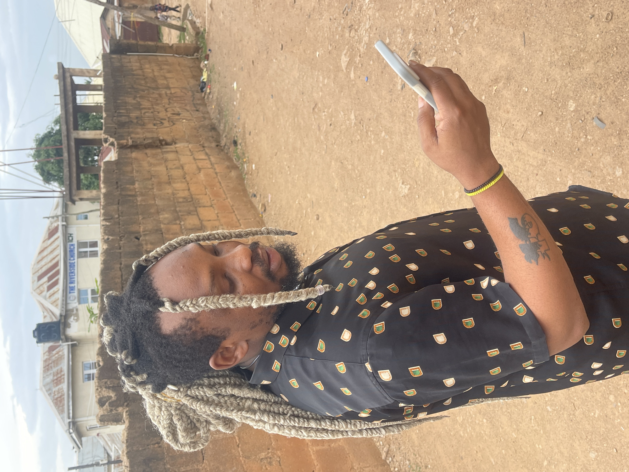 A man with blonde dreadlocks is standing outdoors, holding a white object that might be a pen or stylus in his right hand. He is wearing a black button-down shirt with an orange and green pattern. A black and yellow bracelet is visible on his right wrist, and there is a tattoo on his right forearm. In the background, there is a building with the words "The Riverside Church ICH" on its facade, suggesting the location is in the city of Jos, Nigeria. The ground appears to be dirt or sand.