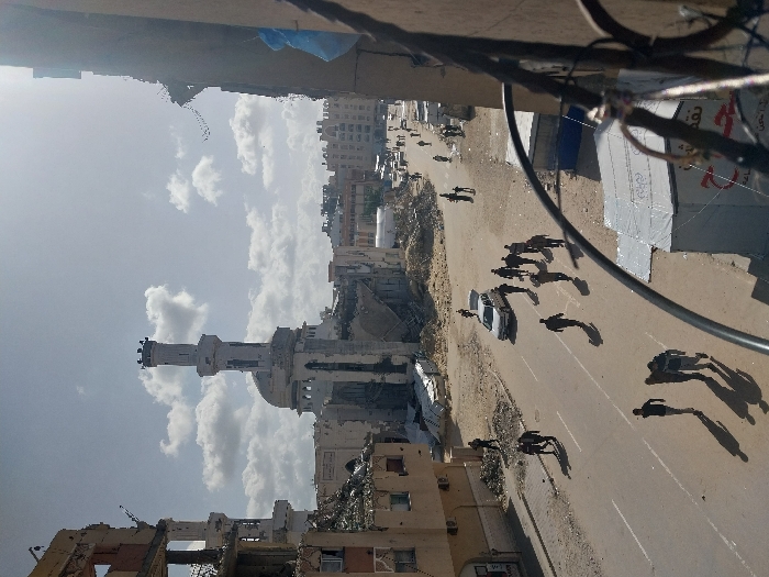 The image depicts a street scene in Khan Yunis, Palestinian Territory, viewed from an elevated perspective, possibly a building balcony, with a metal railing visible in the foreground. A large mosque with a distinct minaret and dome stands prominently in the middle distance, showing significant structural damage, including partial collapse and surrounding rubble. To the left, a multi-story building is heavily damaged, with large sections missing and debris piled at its base.
The paved street below is populated by numerous individuals, approximately 20-30, mostly men, walking in various directions. Most are clustered in groups, casting long shadows. One white sedan is visible on the street, with its trunk open, and several people are in its vicinity. The ground surrounding the street and damaged structures is covered in sand, dirt, and scattered debris.
Further down the street, more damaged buildings and piles of rubble are discernible. Towards the bottom right, next to the street, there are makeshift structures or tents constructed from tarpaulin, one bearing legible Arabic text. The sky is partially cloudy with bright sunlight, illuminating the scene and creating strong shadows. The overall environment conveys widespread destruction and ongoing human activity amidst the rubble.